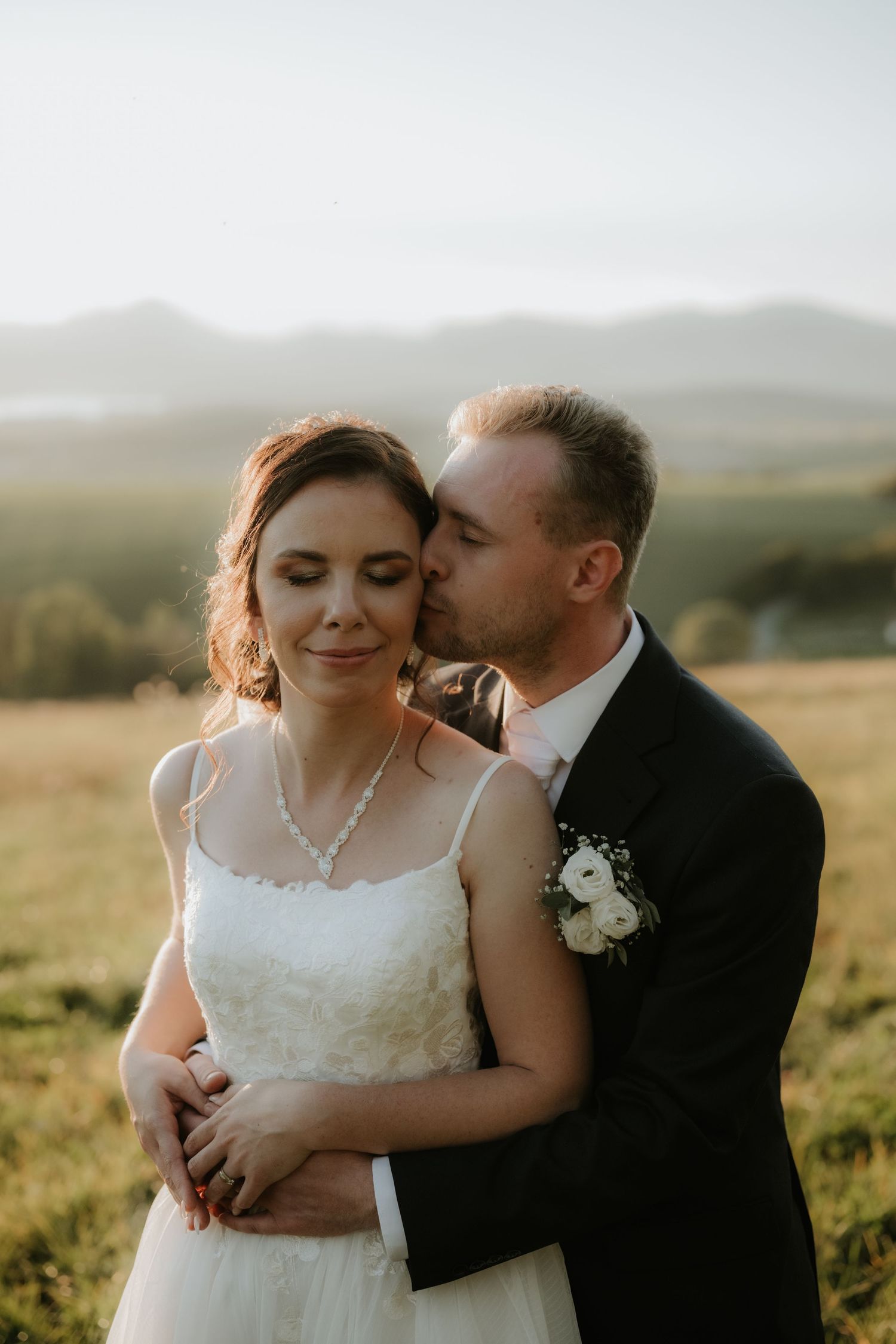 Groom in black suit kisses bride in white lace dress outdoors at golden hour with mountain landscape backdrop.
