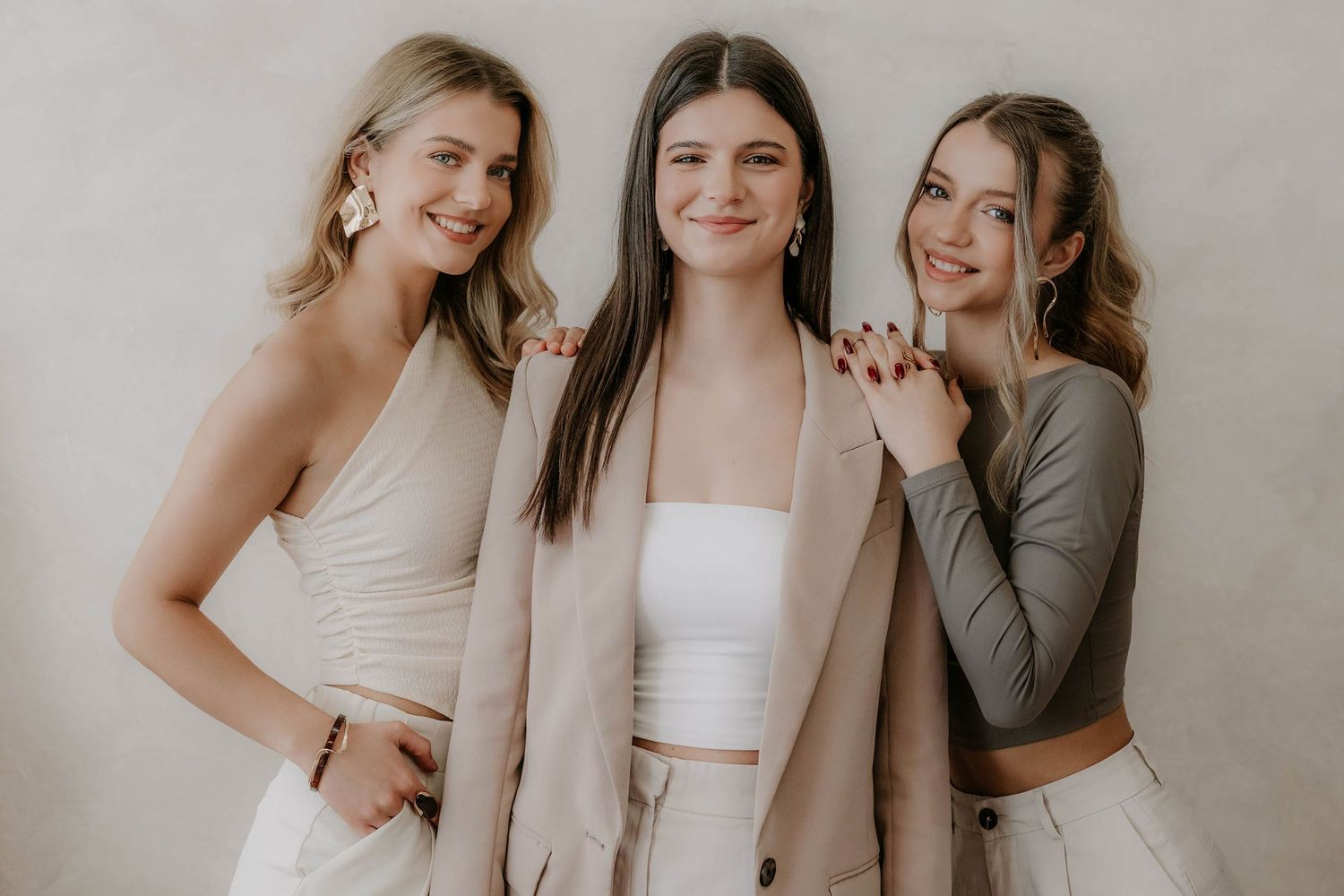 Three stylish young women posing together in neutral toned outfits against beige wall.