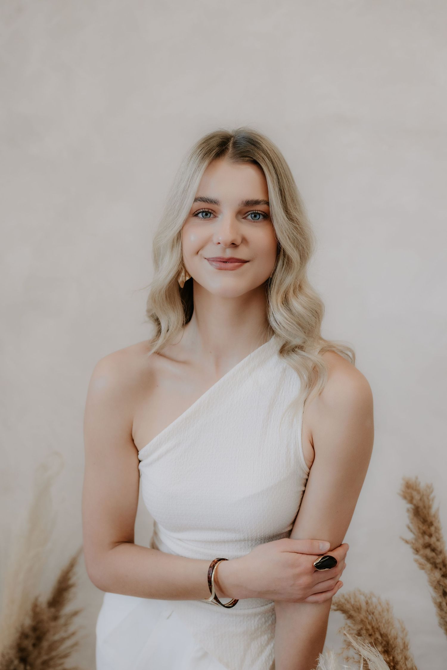 Elegant blonde woman in white one-shoulder dress posing against a neutral beige background.