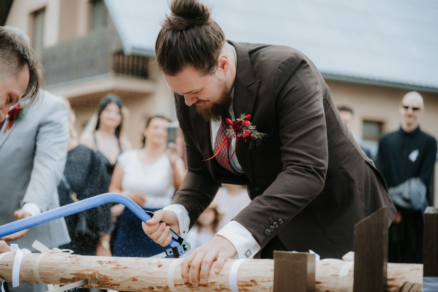 Groom in brown suit sawing wood log at outdoor wedding ceremony with guests watching in background.