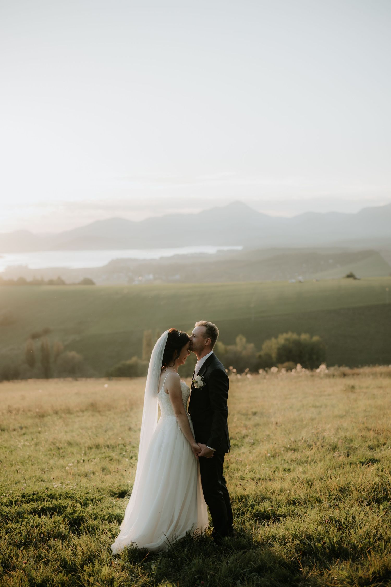 Bride and groom sharing a kiss on a green hillside with a scenic mountain and lake vista at golden hour.