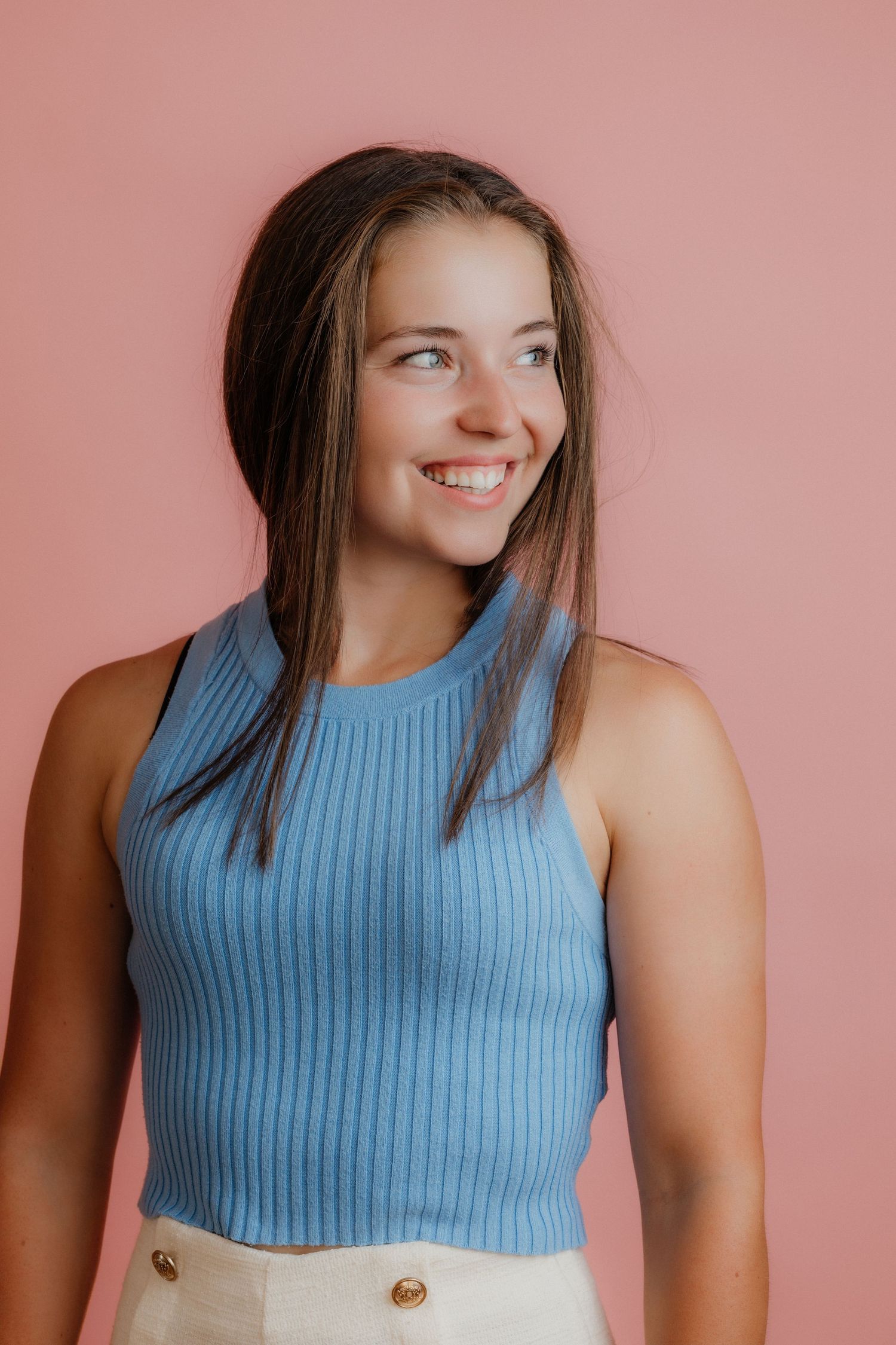 Smiling young woman with long brown hair wearing blue ribbed sleeveless top against pink background.