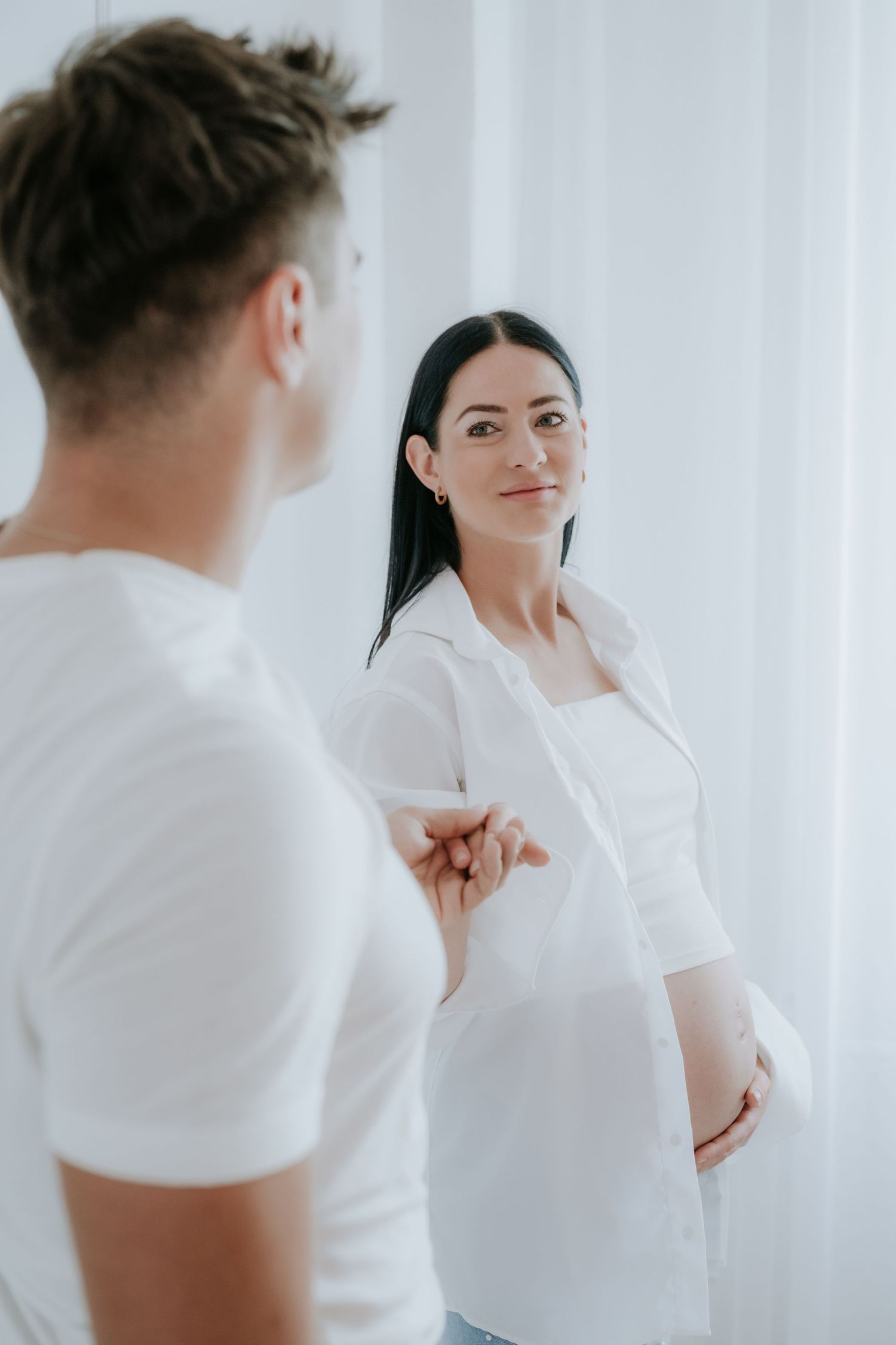 Pregnant woman in white shirt holds partners hand in bright airy white studio setting.