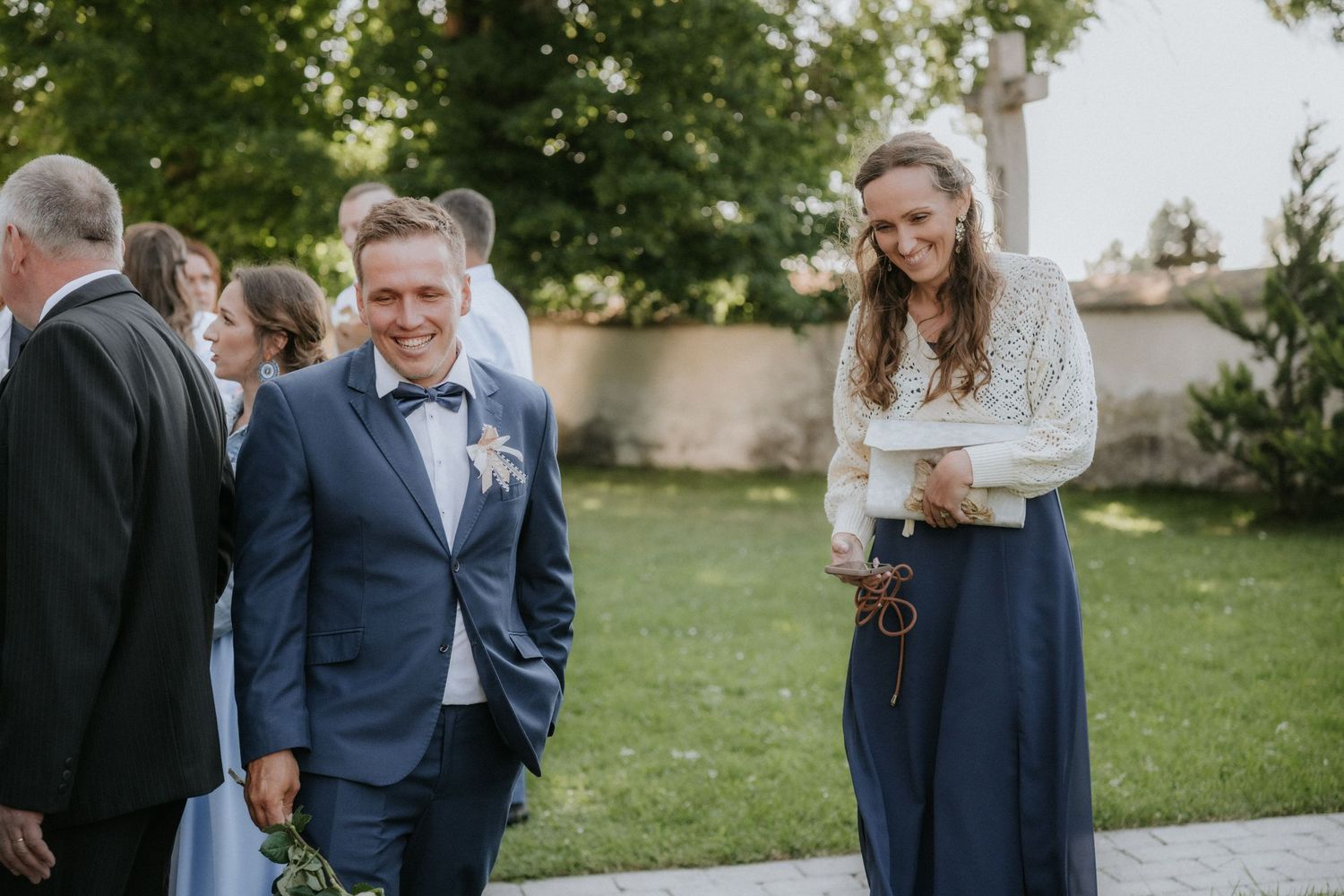 Smiling groom in blue suit with boutonniere walks with a woman in white lace top and navy skirt at outdoor wedding.