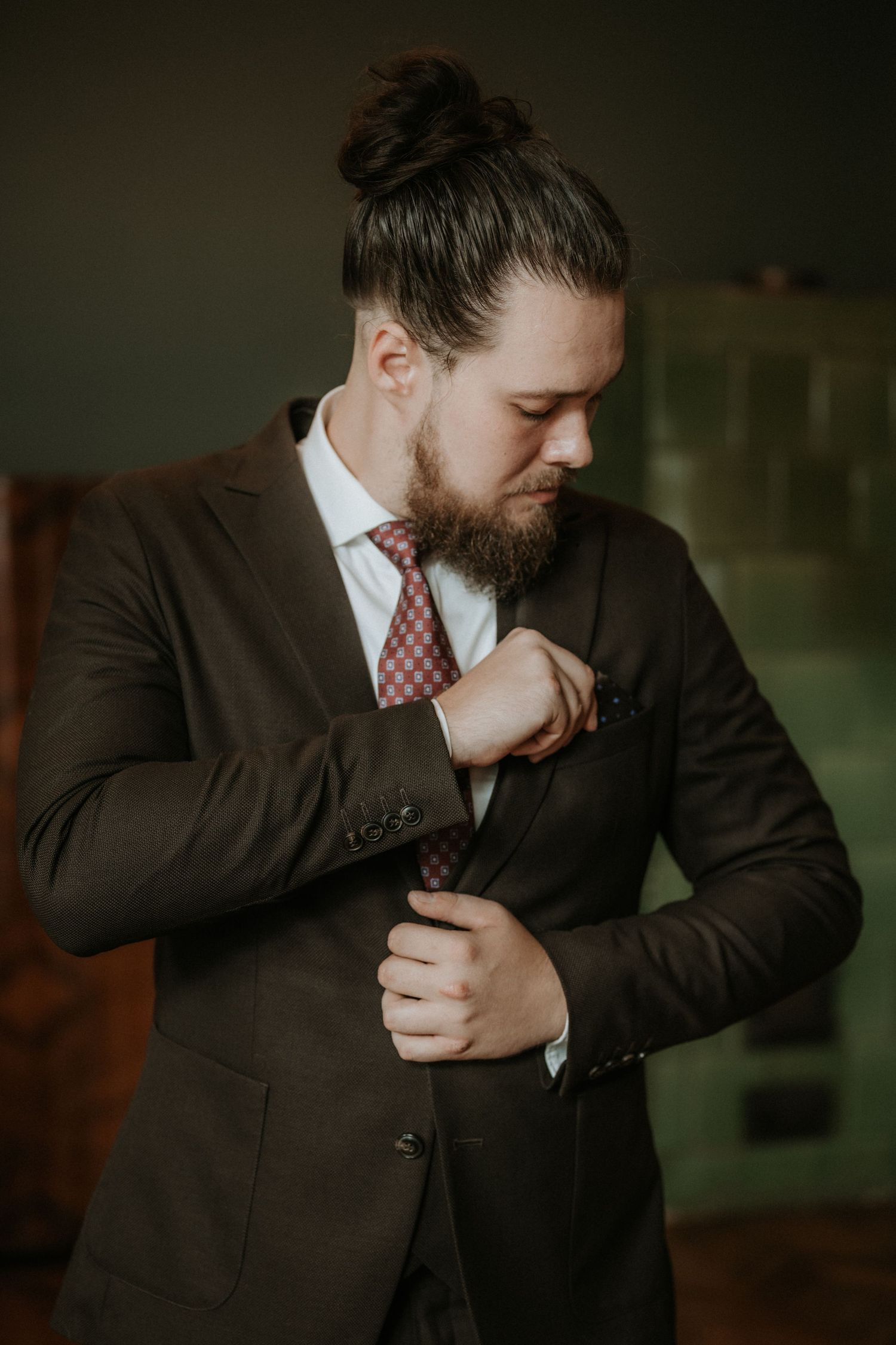 Bearded man with hair bun adjusting his tie, wearing a dark suit and red patterned tie in a dimly lit room.