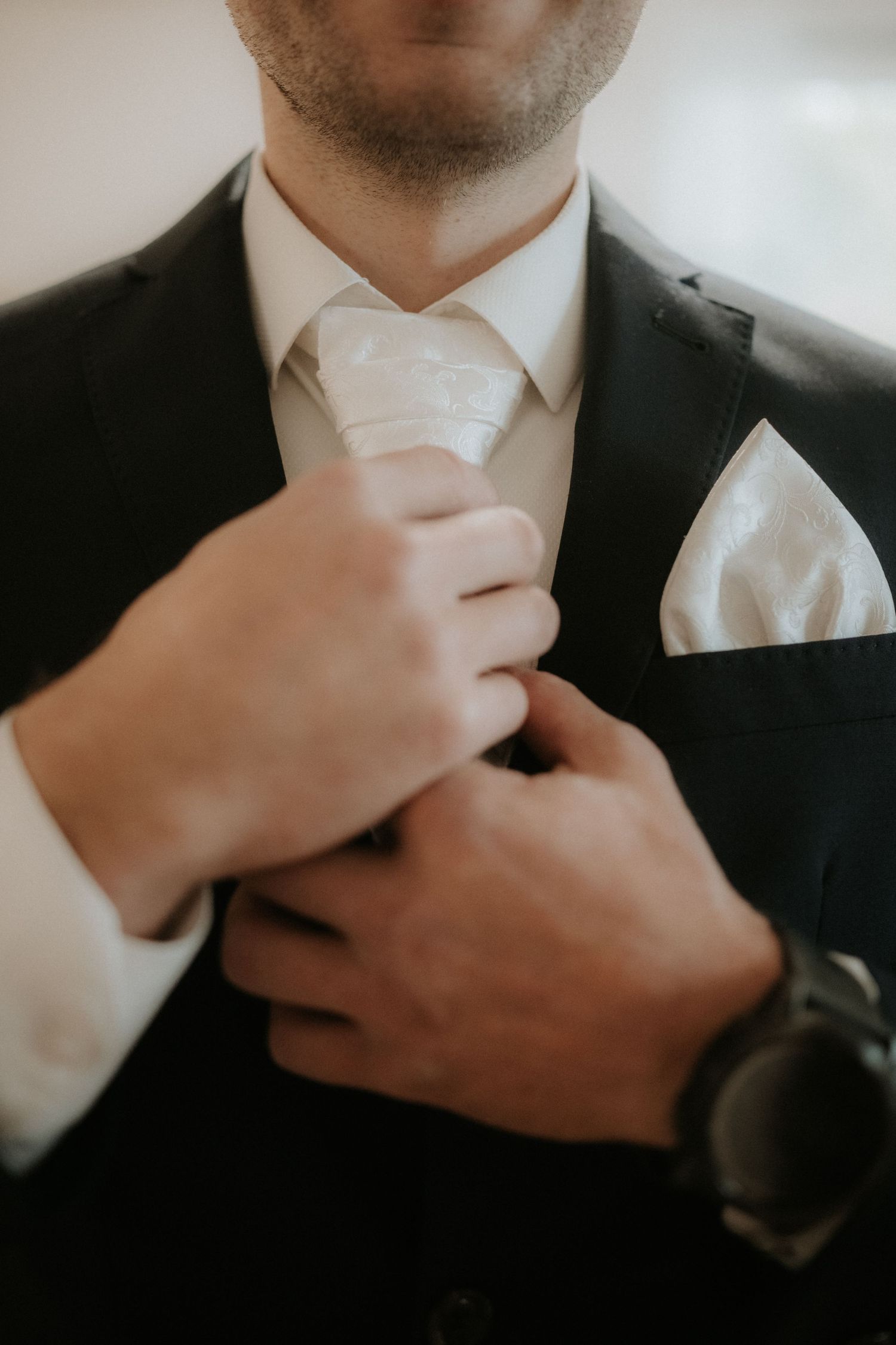 Groom in black suit adjusting white paisley tie with matching pocket square, wearing a watch.
