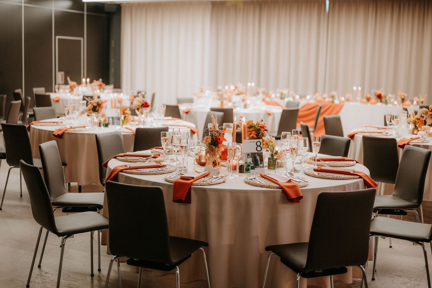 Elegant banquet hall with round tables set for a wedding reception featuring orange napkins and floral centerpieces.
