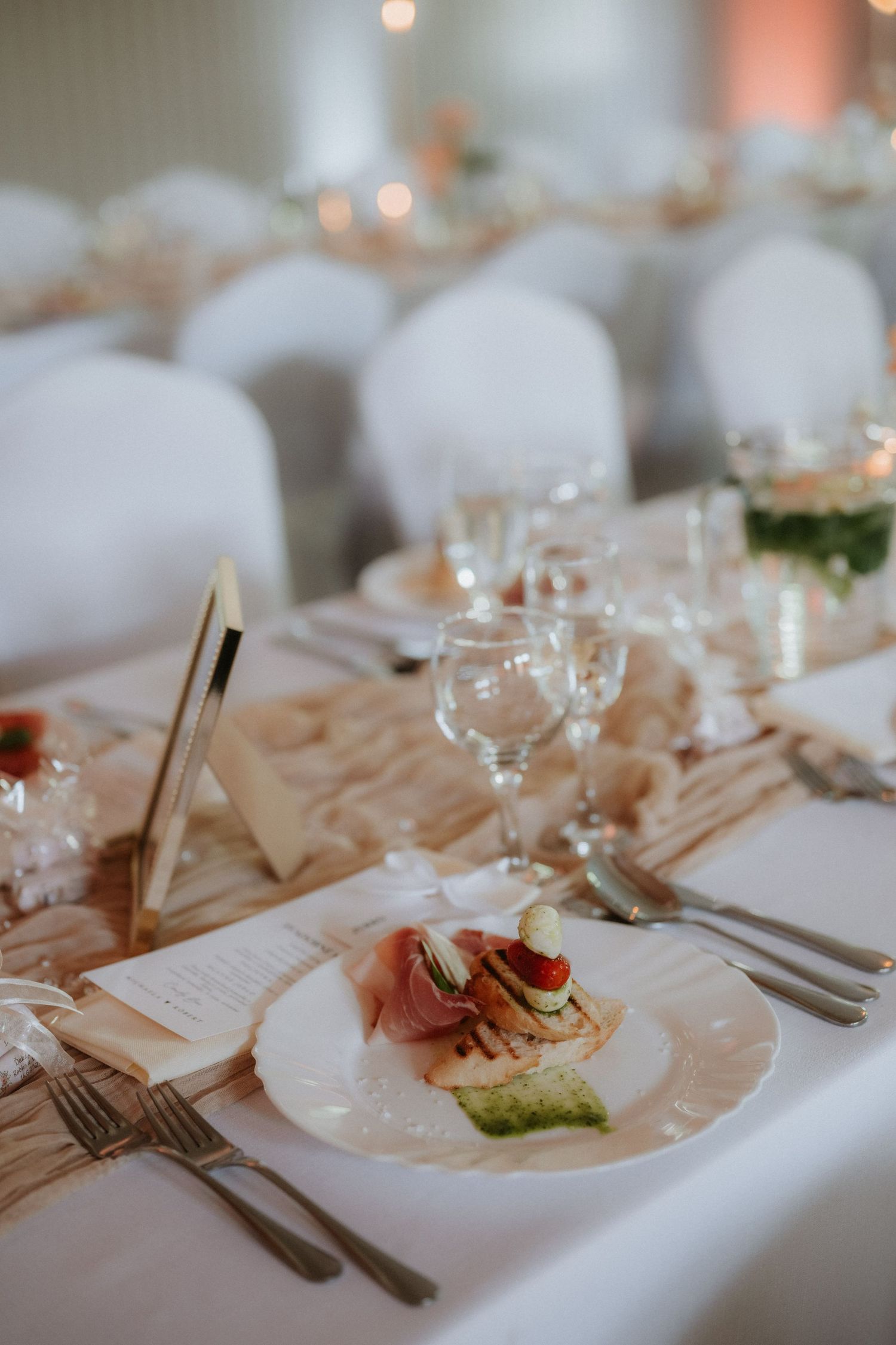 Elegant wedding reception table setting with white plates, crystal glasses, silverware, and appetizers on a white linen tablecloth.