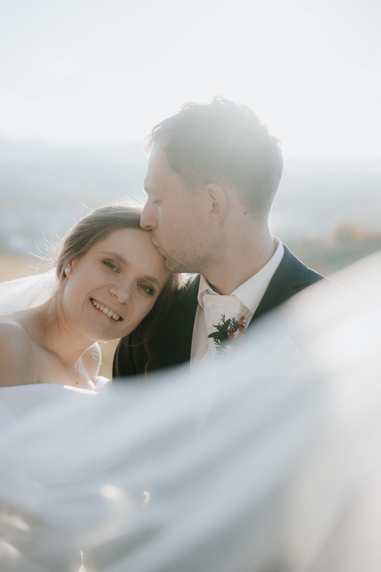 Groom kissing bride on cheek as she smiles, with soft backlight and flowing veil in foreground at outdoor wedding.