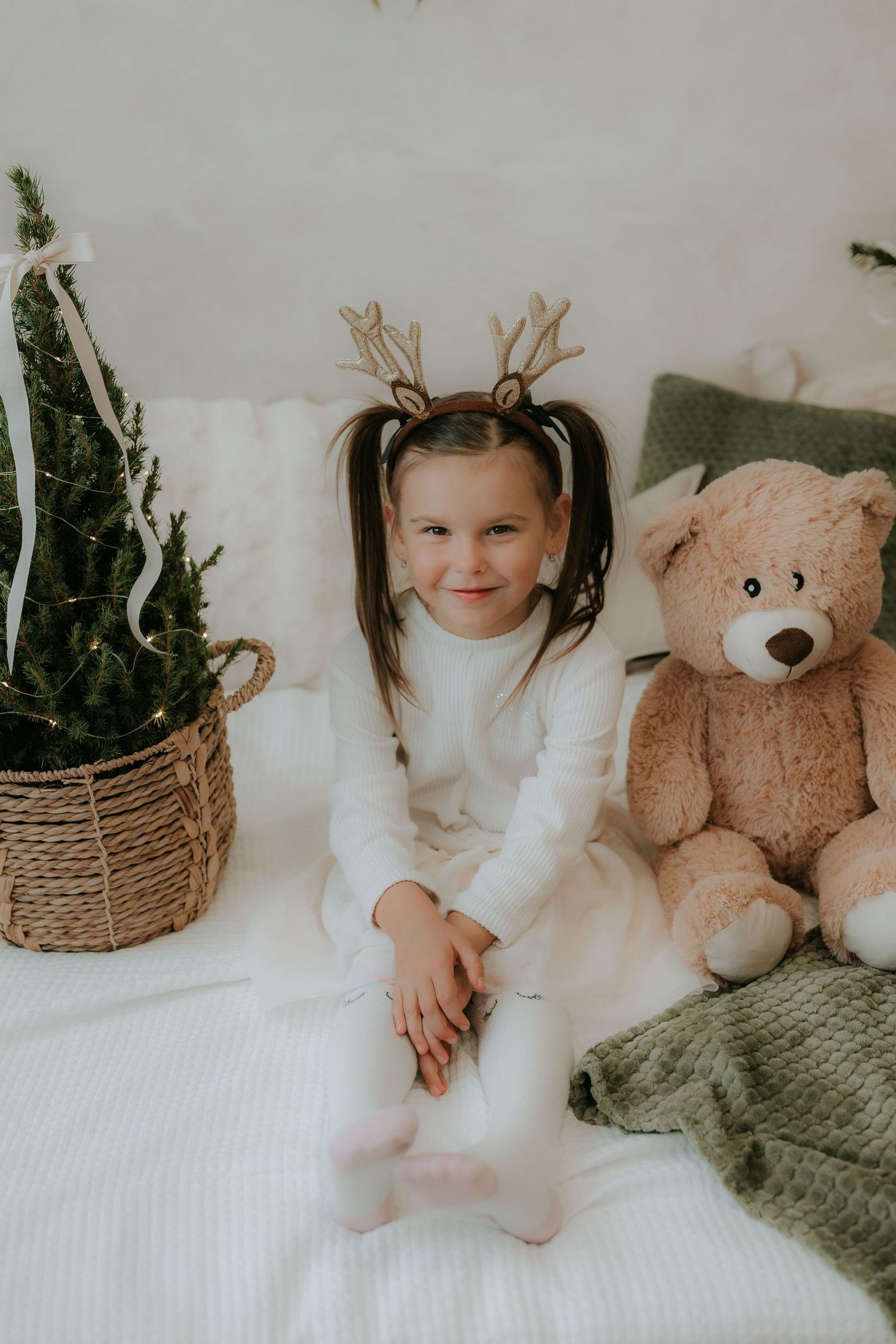 Young girl in white outfit with reindeer antler headband sits on bed with teddy bear and Christmas tree.