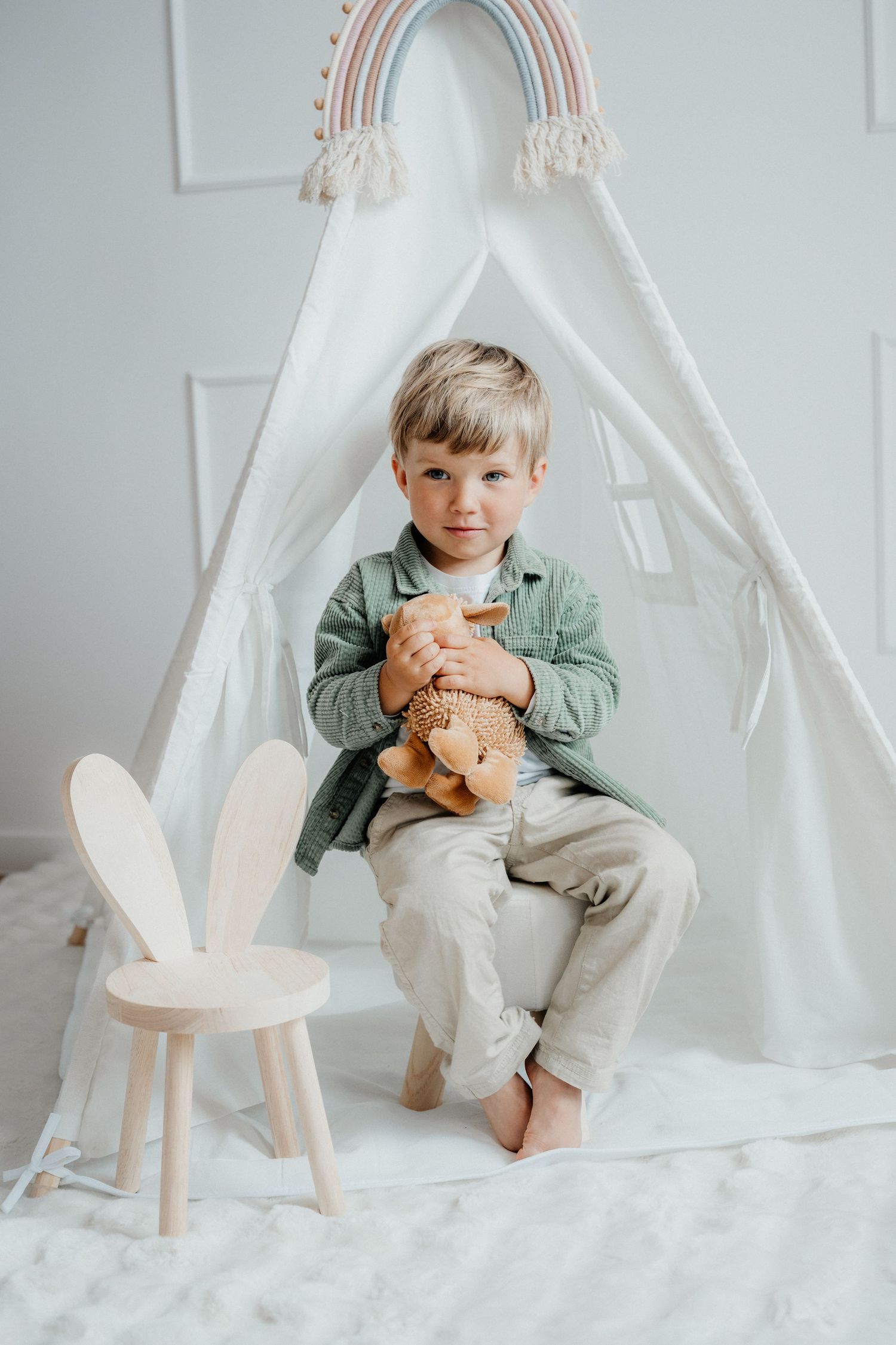 A smiling toddler boy in a green shirt holds a stuffed animal while sitting by a white play tent.