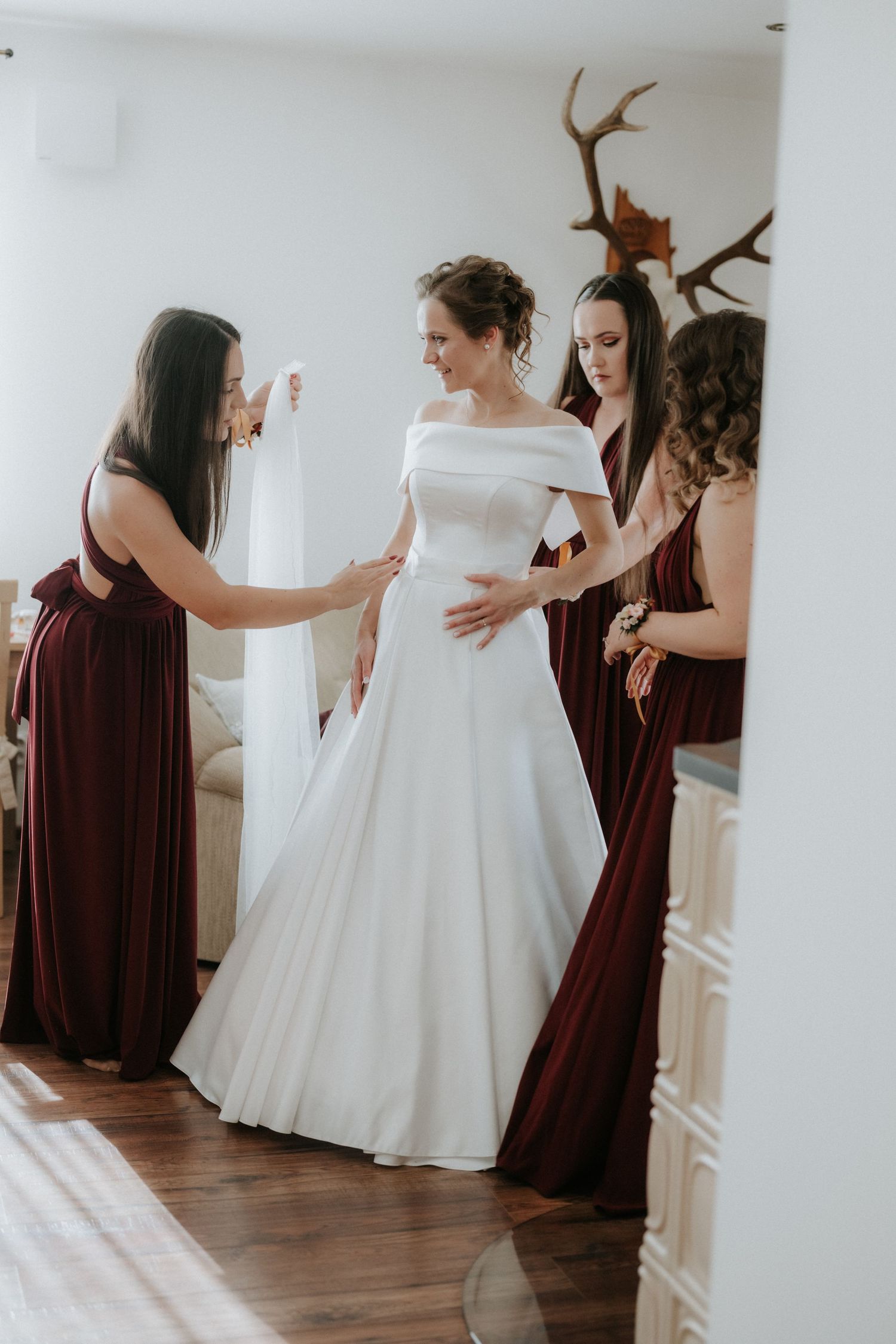 Bride in white off-shoulder gown assisted by two bridesmaids in burgundy dresses during wedding preparation.