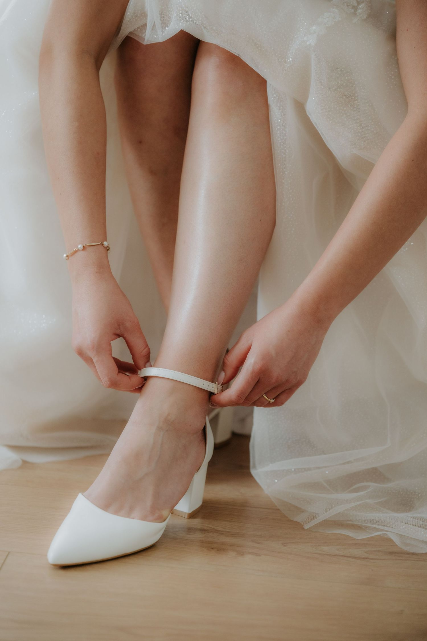 Bride fastening white block heel ankle strap wedding shoes while wearing a flowing white bridal gown.