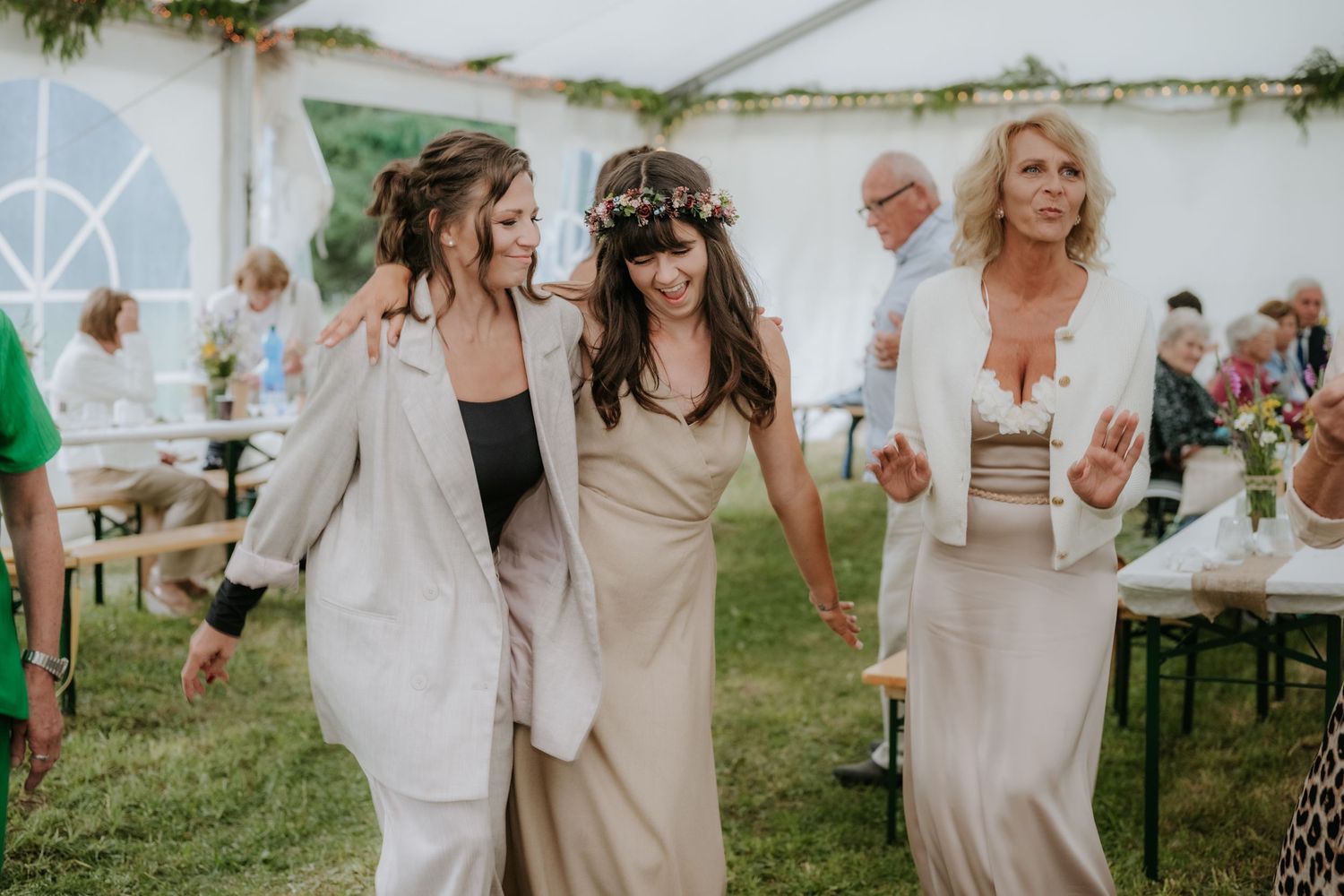 Three women in elegant attire walk together under a white tent at an outdoor wedding celebration.