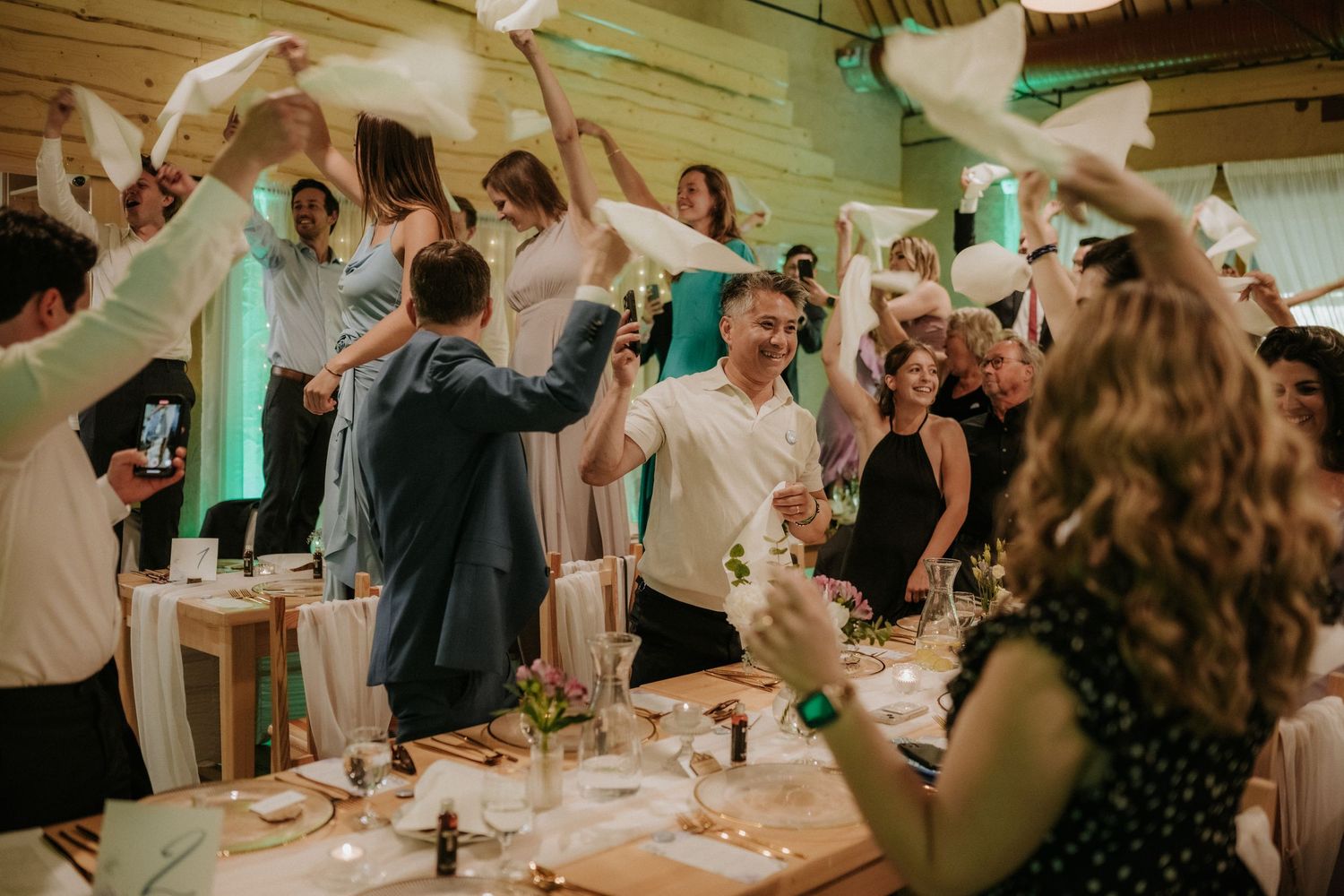 Guests celebrate at a wedding reception, waving white napkins in a festive indoor venue with wooden walls.