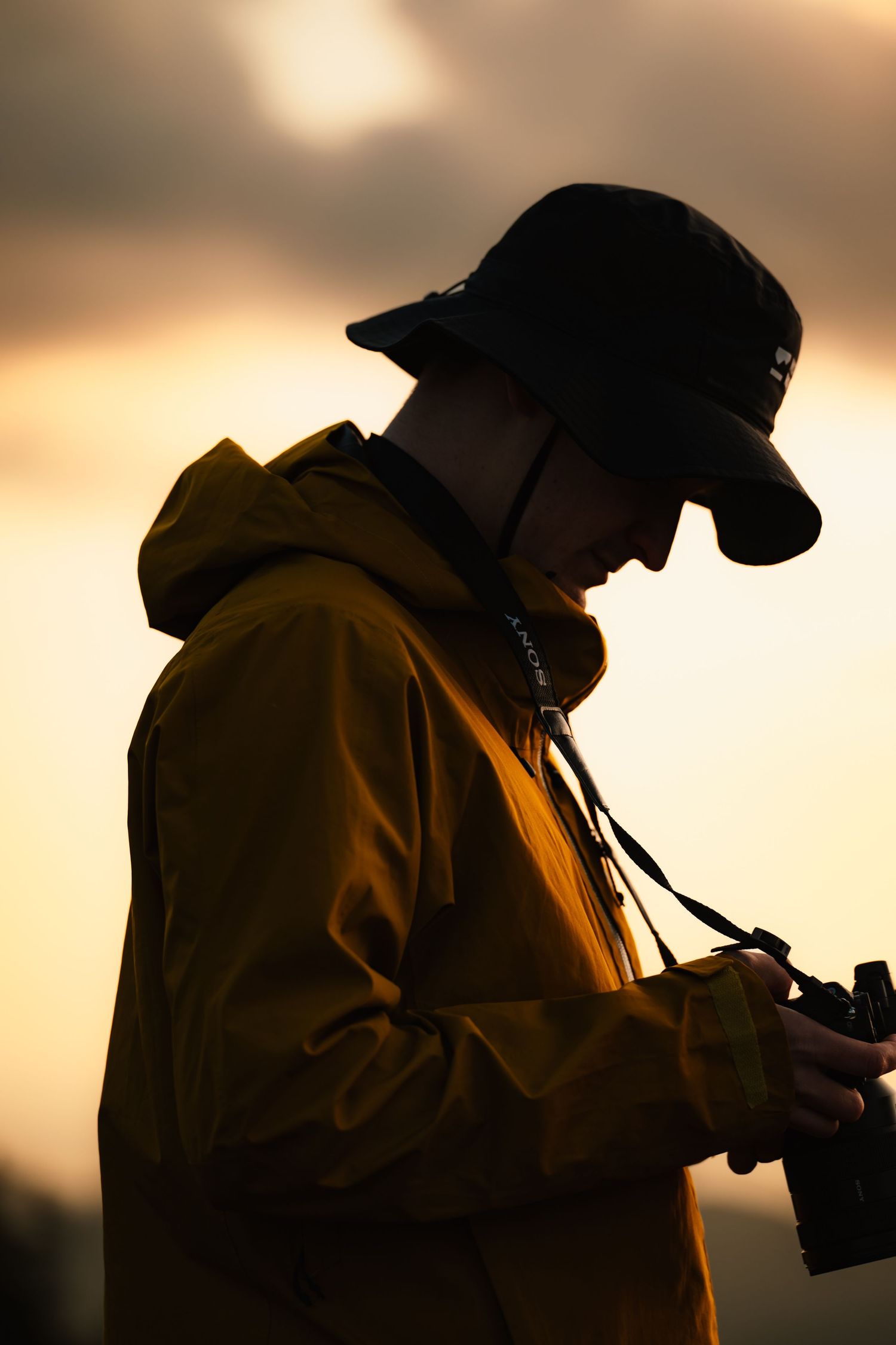 Silhouette of a photographer in a yellow jacket and bucket hat reviewing images at golden sunset.