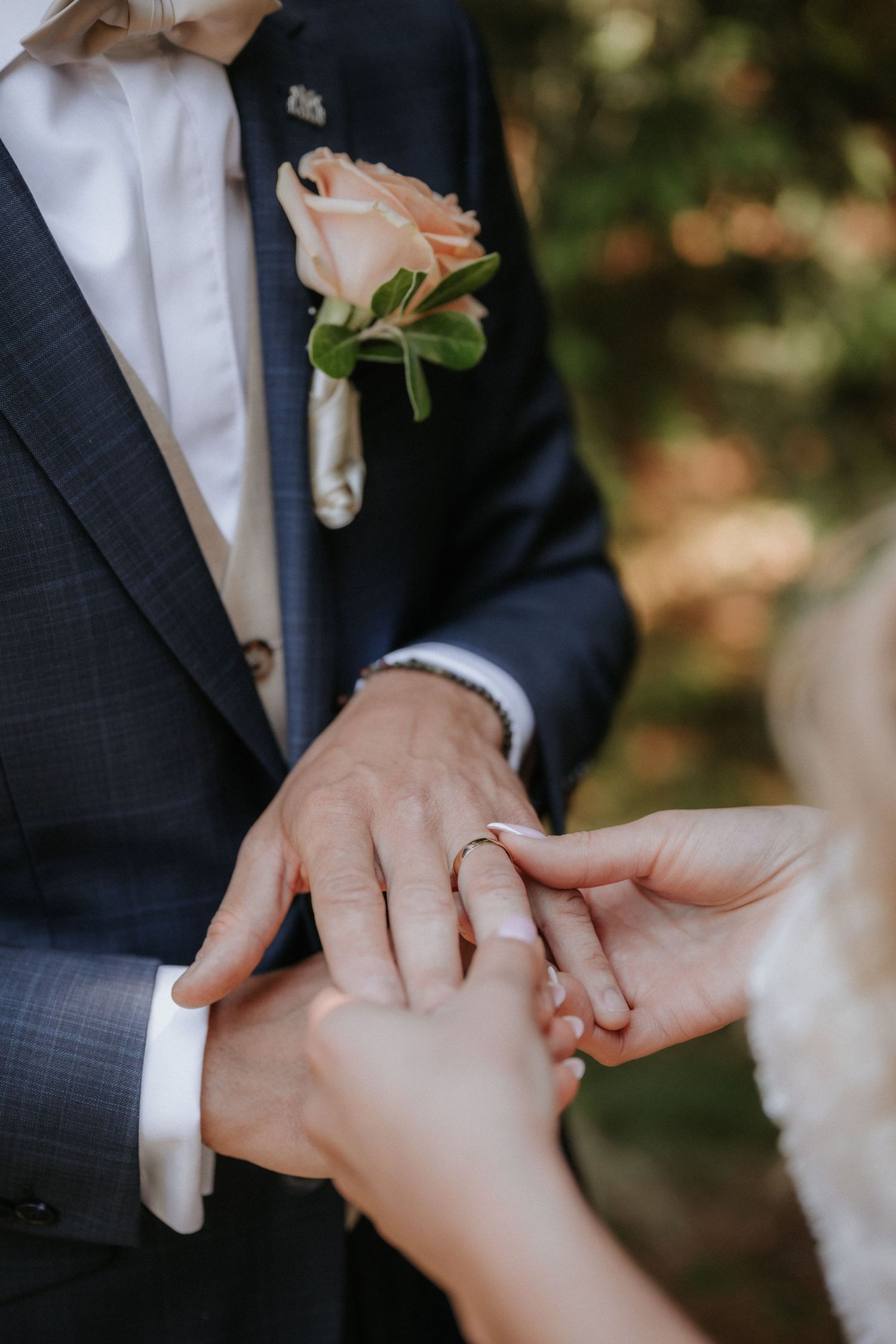 Bride placing a gold wedding ring on groom's finger, groom wearing navy suit with peach rose boutonniere.