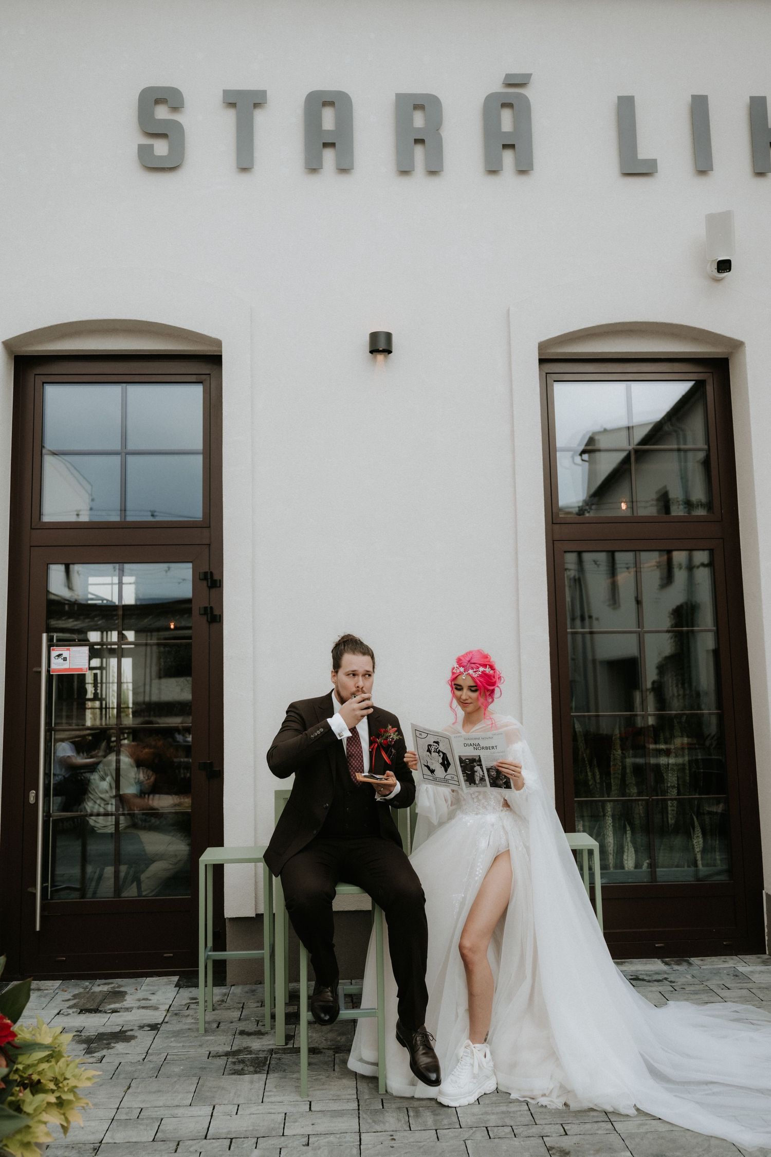 Bride in white gown with pink hair and groom in suit sitting outside Stara Li venue, sharing drinks.