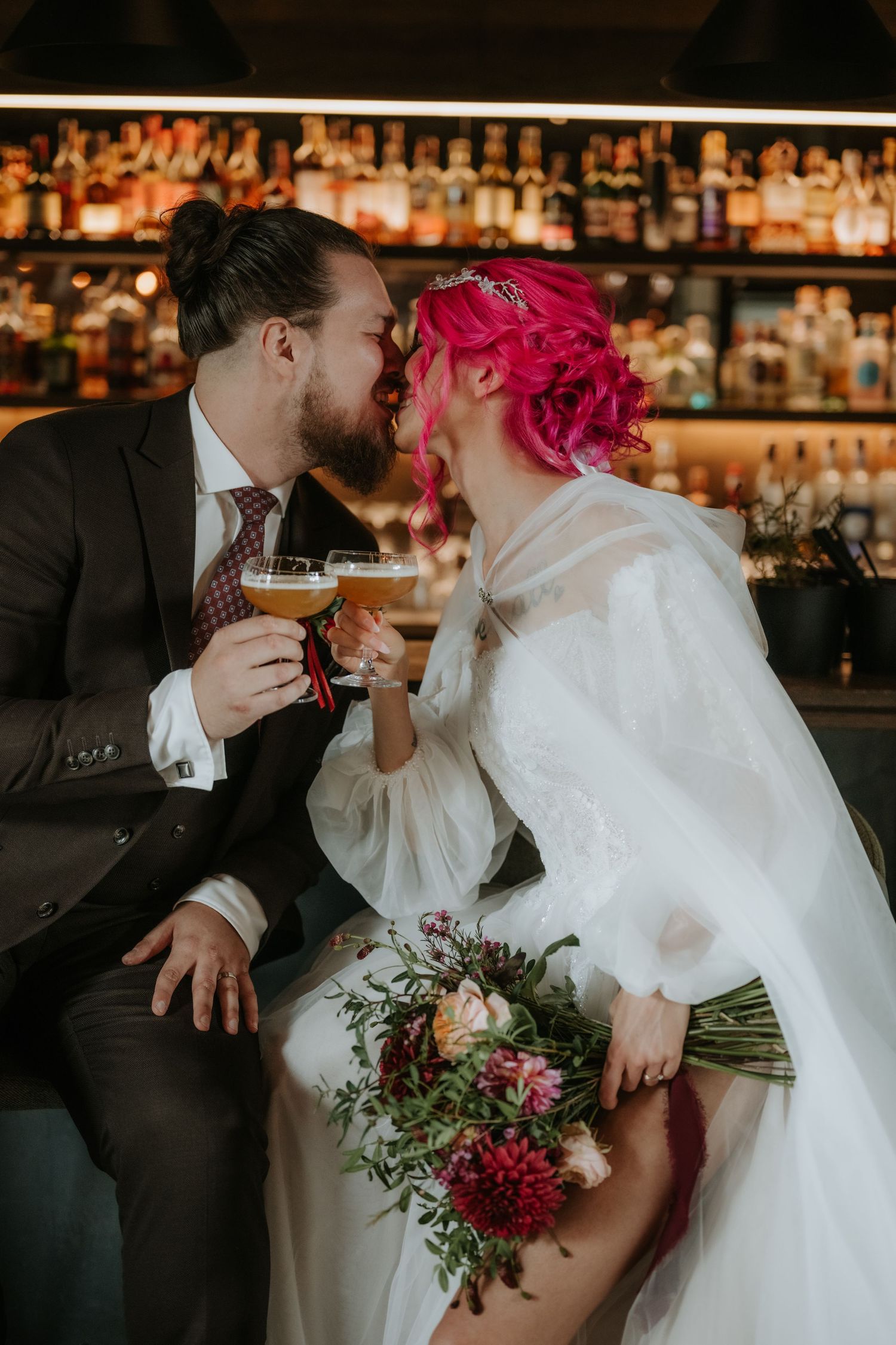 Newlyweds kissing and toasting with cocktails at a bar, bride with pink hair holding a floral bouquet in a white gown.