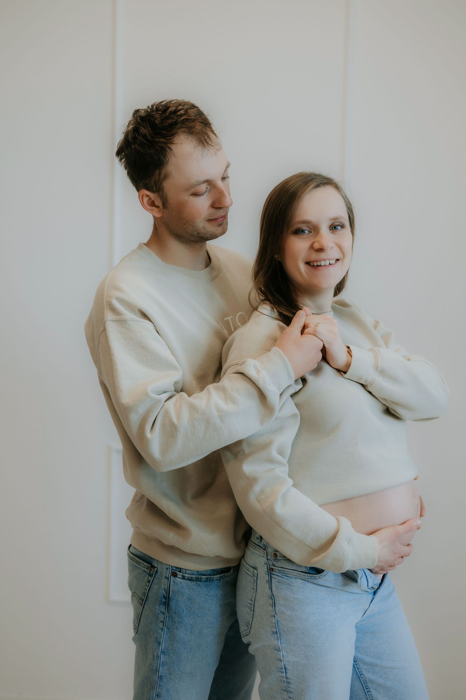 A smiling pregnant couple in matching beige sweatshirts and jeans embrace tenderly in a bright studio.