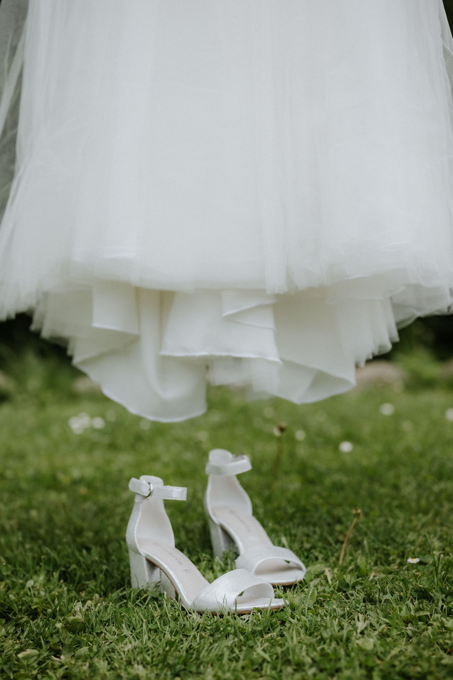 White bridal heeled sandals on green grass beneath a flowing white tulle wedding dress hem.