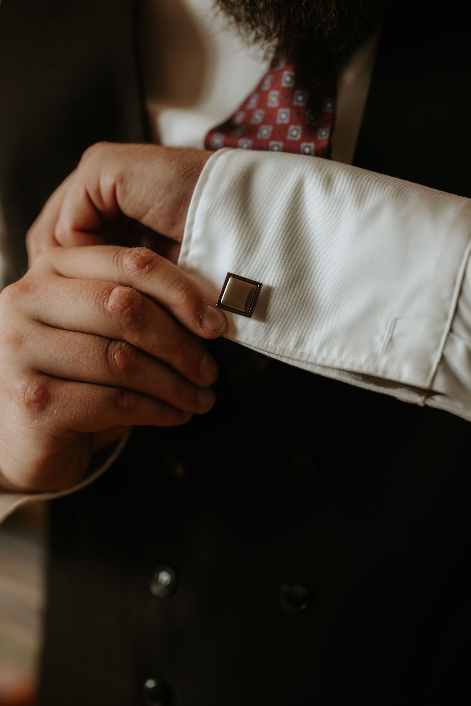 A man in a dark suit adjusts his silver cufflink on a white French cuff shirt, wearing a red patterned tie.