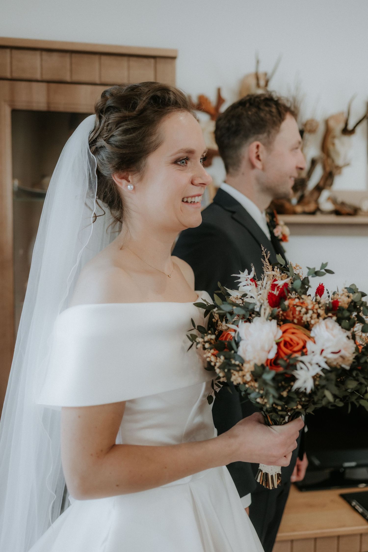 Smiling bride in off-shoulder white gown holding autumn bouquet with orange roses, groom in dark suit behind her.