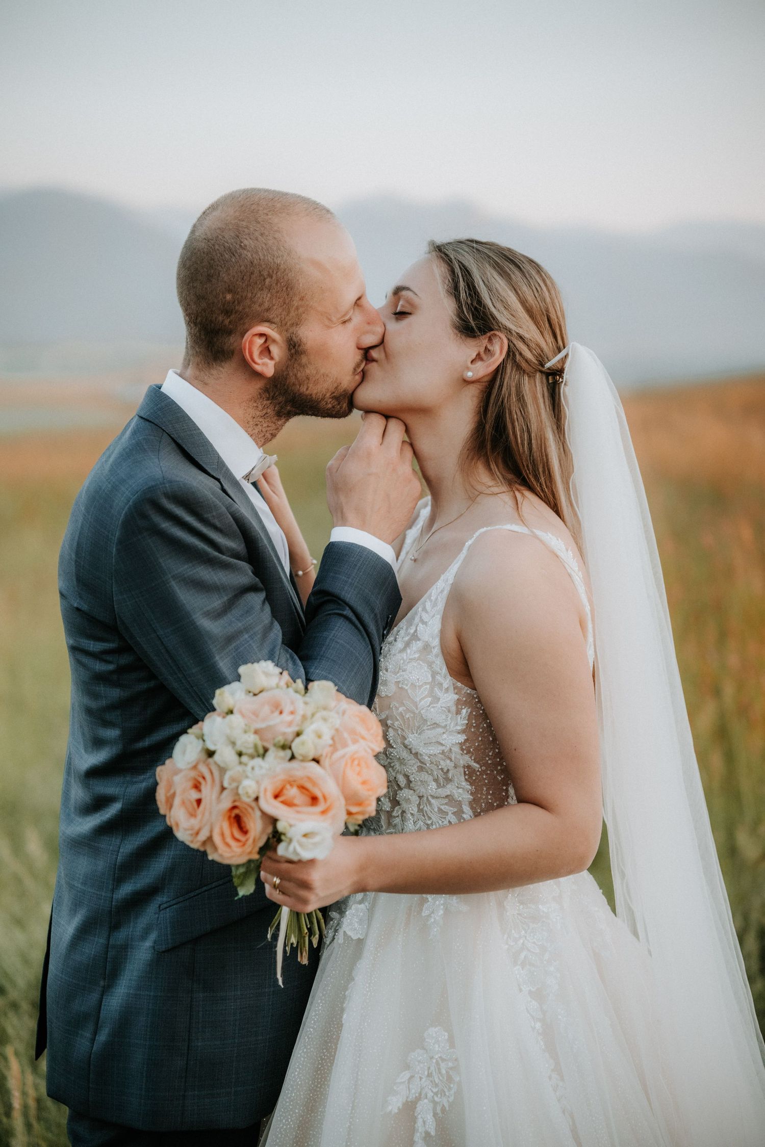 A bride and groom share a romantic kiss in a golden field, she holds a peach rose bouquet while wearing a lace gown and veil.