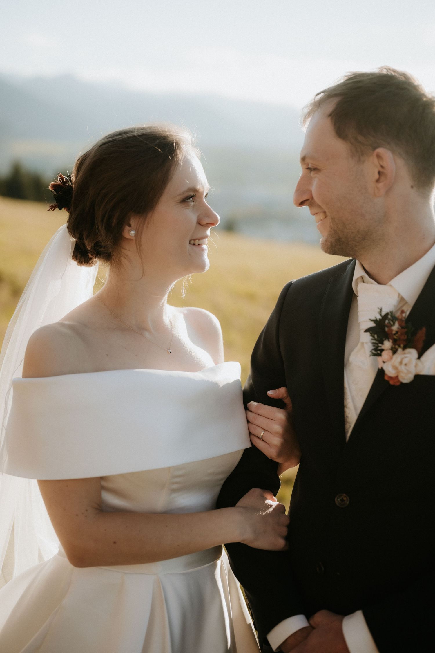 Bride in off-shoulder white gown and groom in black suit smiling at each other in a sunny field.