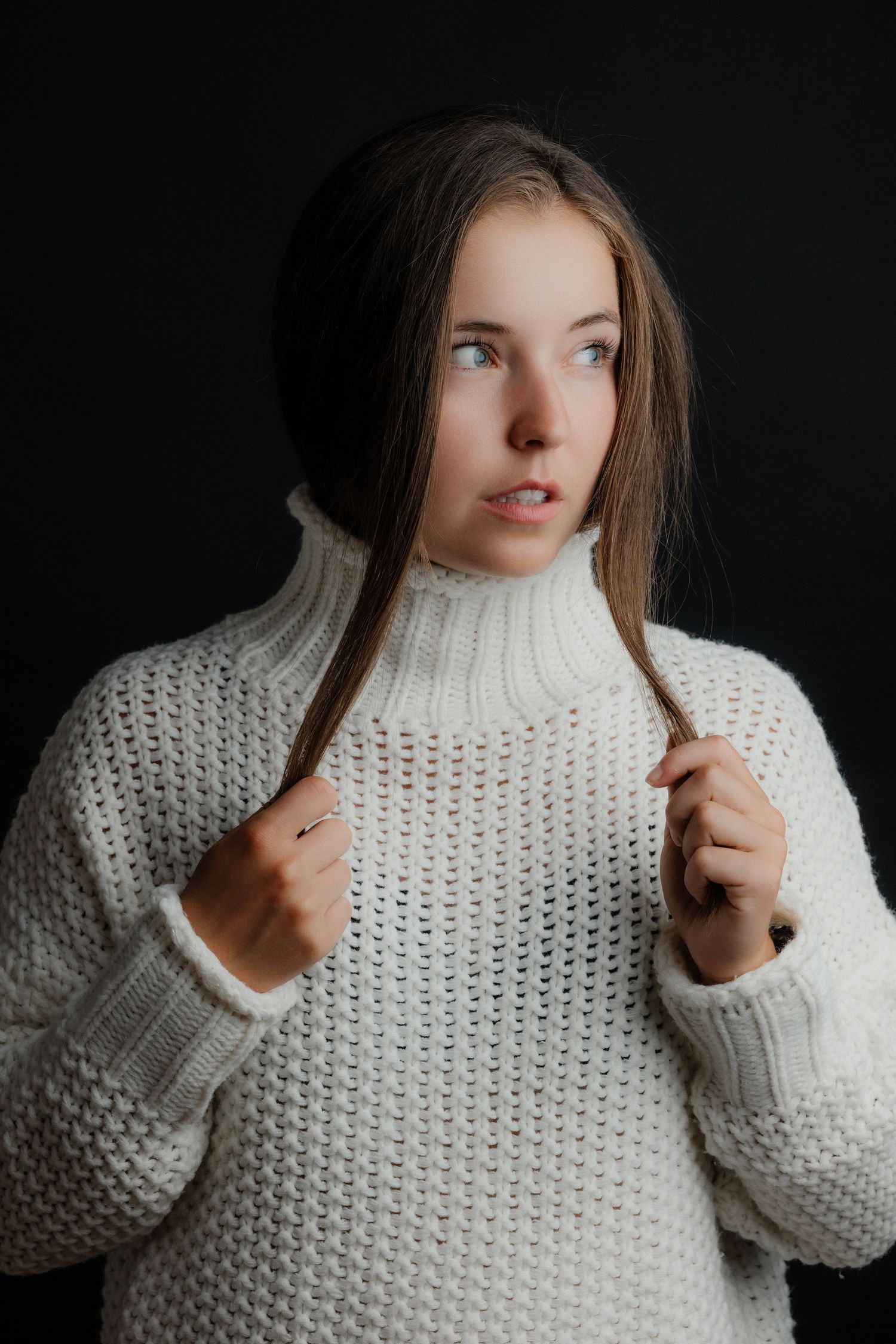 Young woman in a white chunky knit turtleneck sweater holding her hair against a black background.