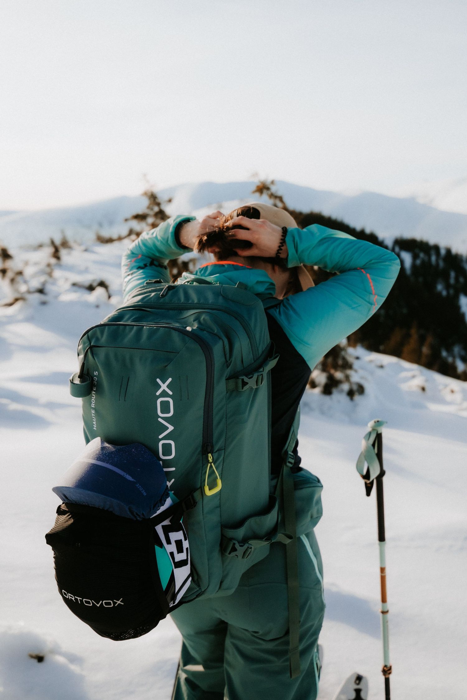 Backcountry skier adjusting helmet with Ortovox backpack on a snow-covered mountain landscape.