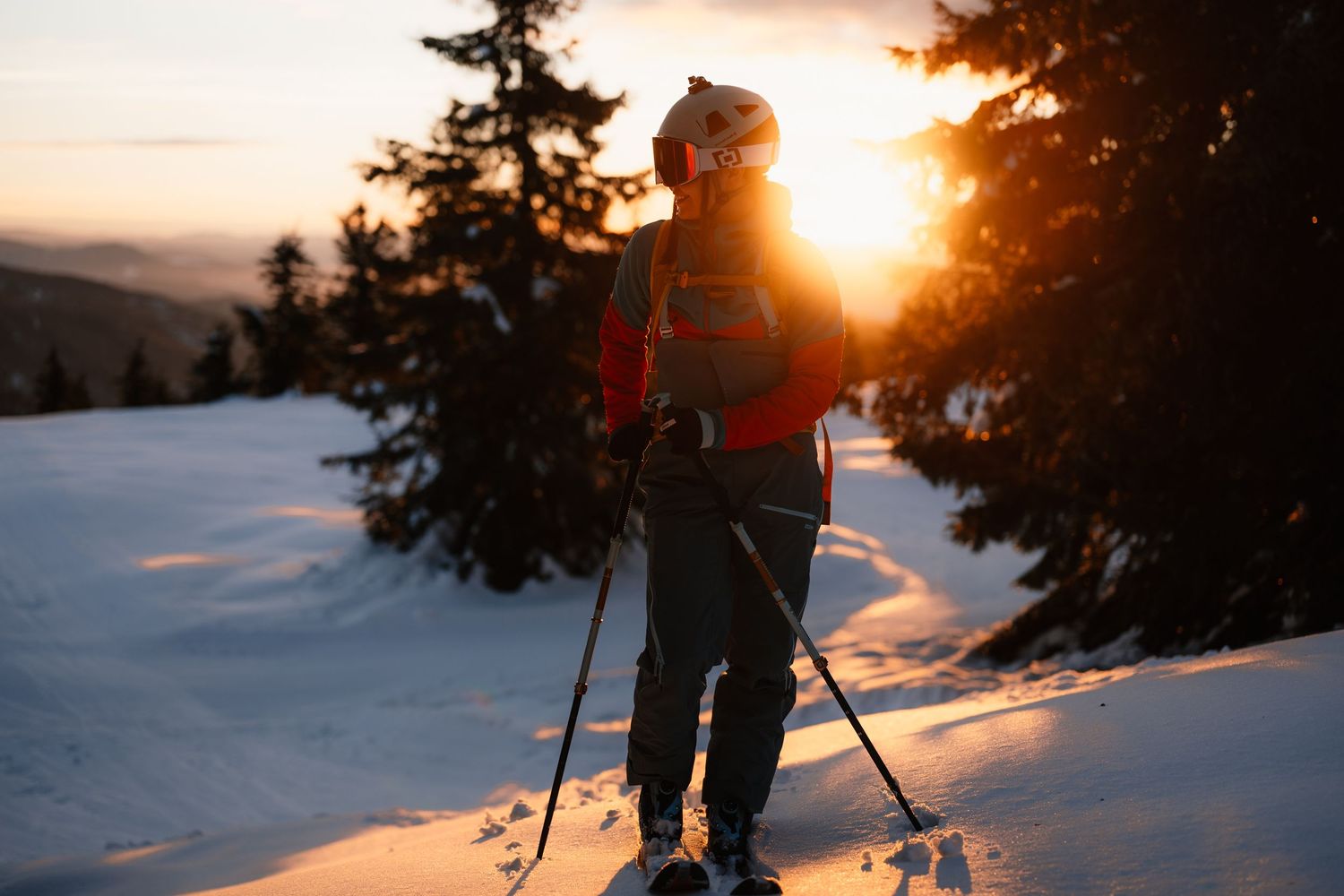 Skier silhouetted against glowing orange sunset light on snow-covered mountain with pine trees.