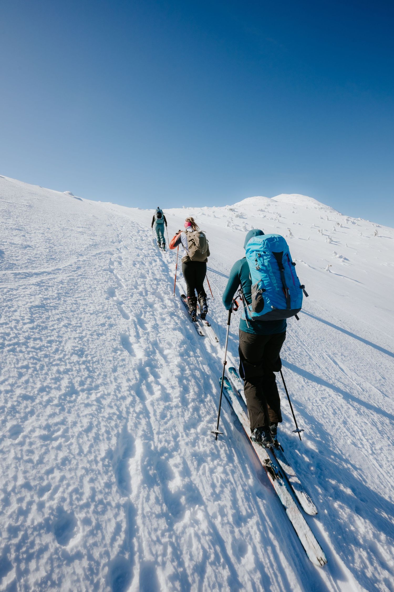 Group of three skiers with backpacks ski touring up a pristine snowy mountain slope under blue sky.