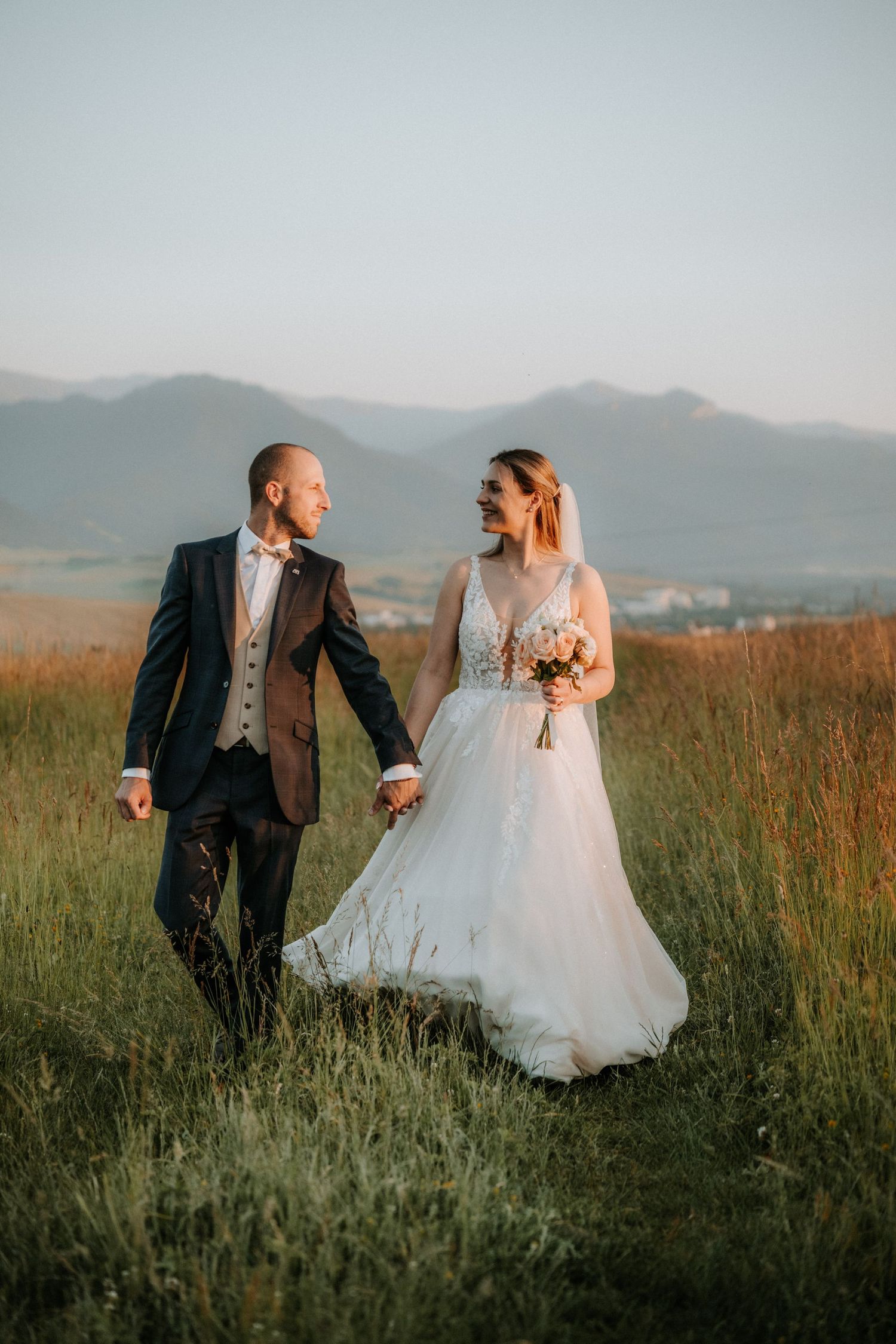 Bride and groom walking hand in hand through a green meadow with mountain backdrop during golden hour sunset.