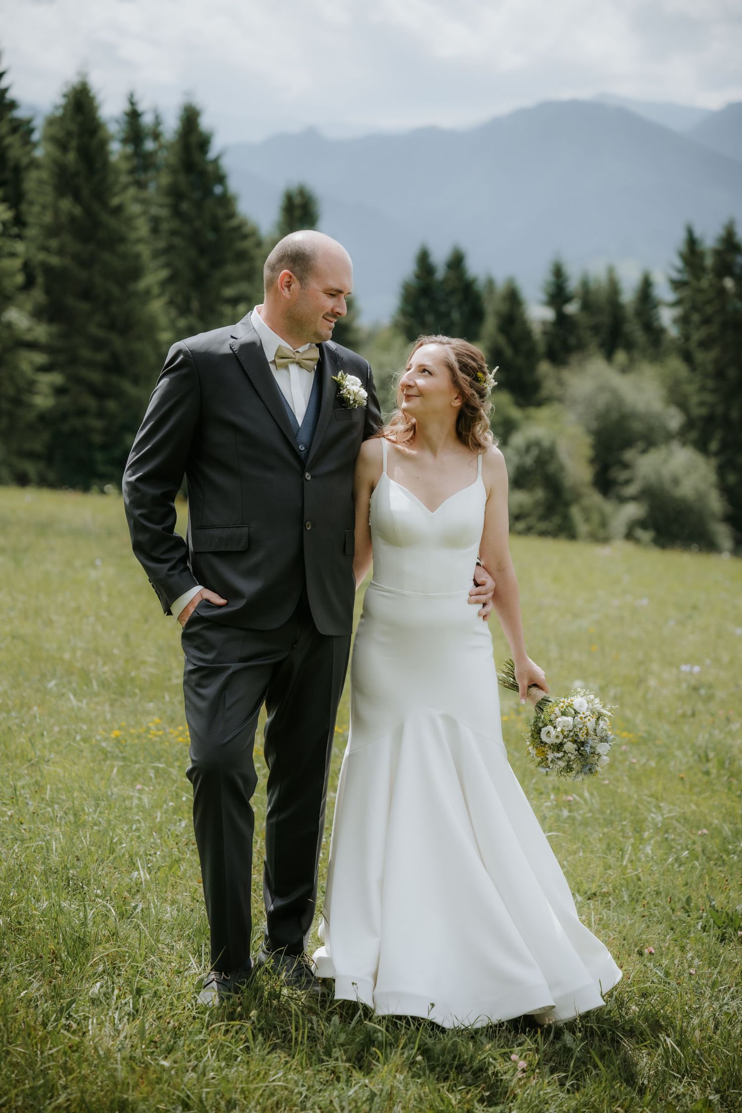 A bride and groom pose together in a green alpine meadow, she holds a white bouquet, mountains and pine trees in the background.