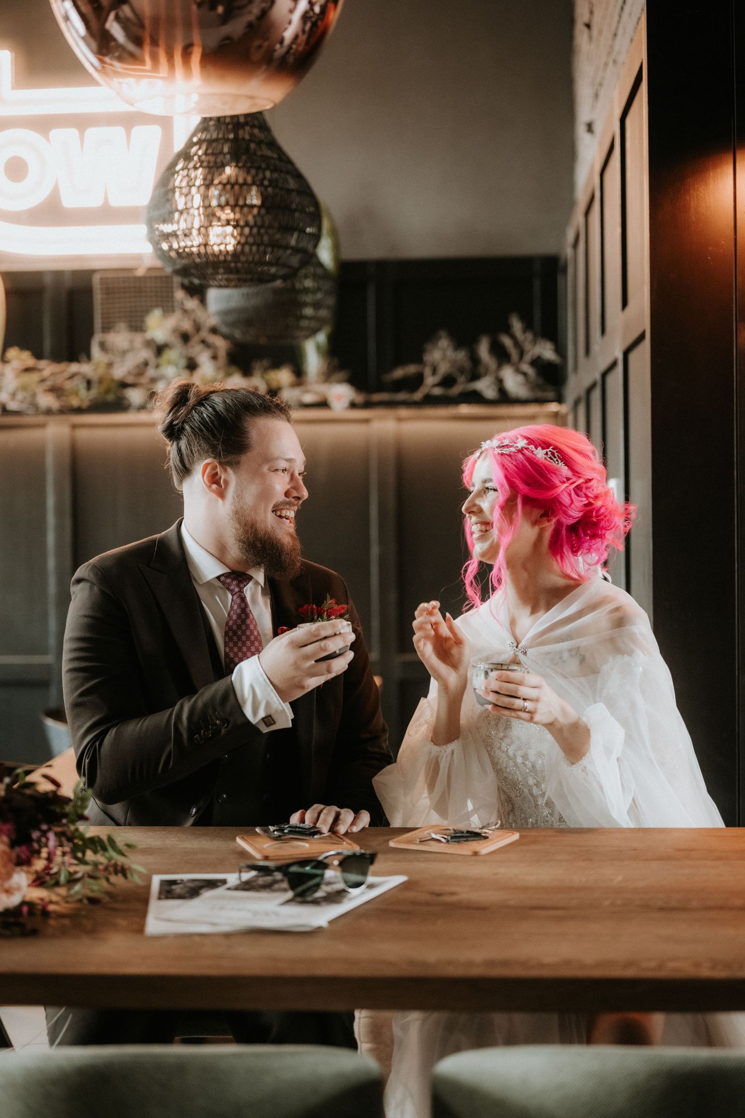 Bride with pink hair and groom in dark suit share a joyful moment at a wooden table in a stylish venue.