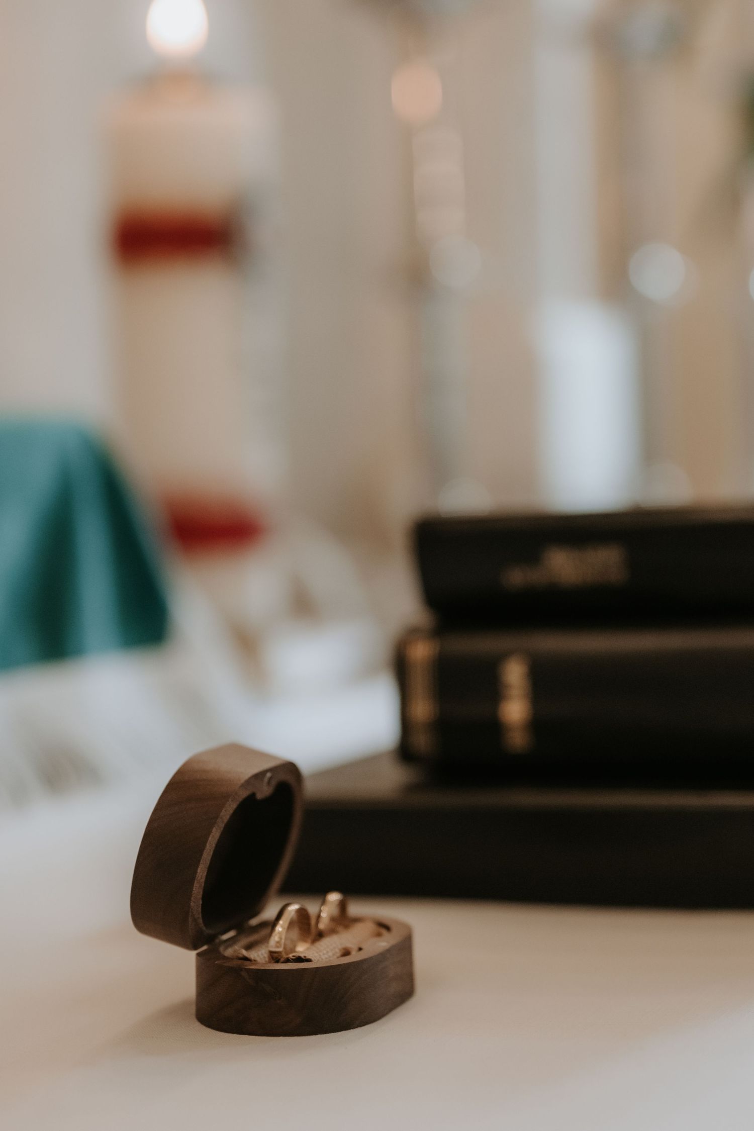 Wedding rings in a wooden box beside stacked black Bibles on a white table, with candles softly blurred in background.