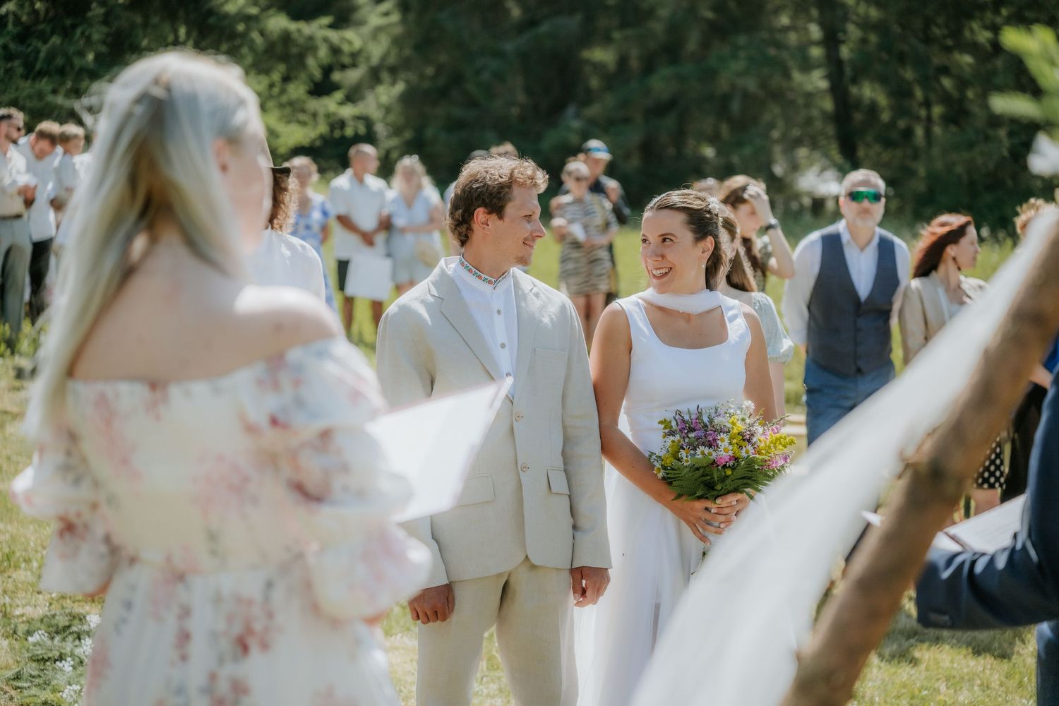 Couple smiling at outdoor wedding ceremony on sunny day, bride holding wildflower bouquet, guests gathered in background.