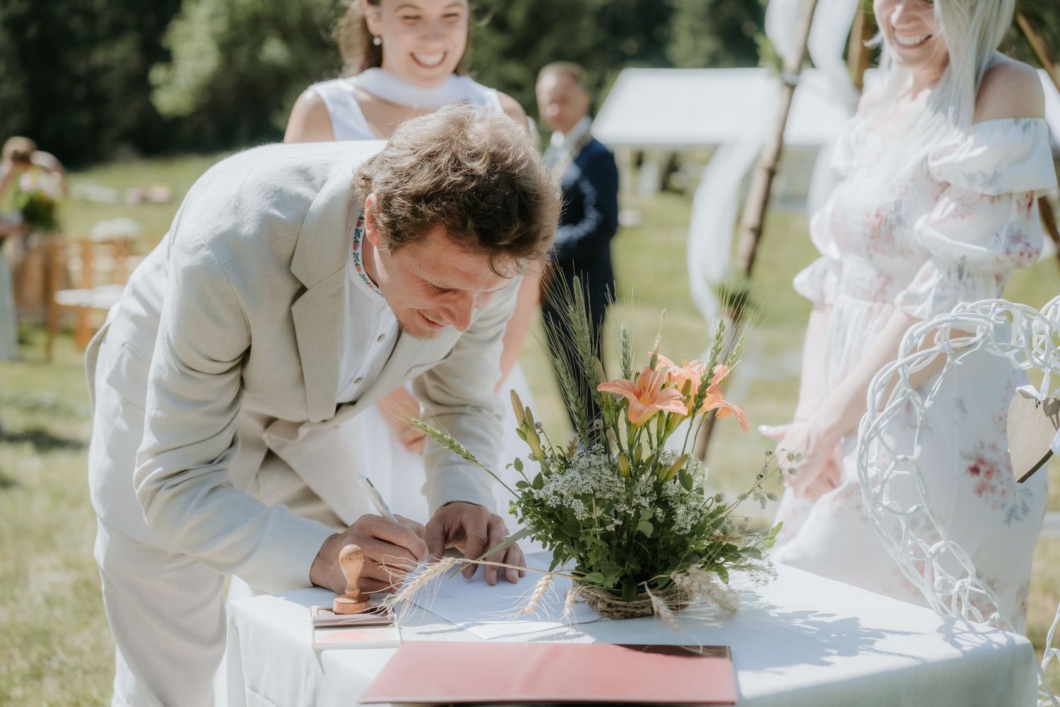 Groom in beige suit signing wedding documents at outdoor ceremony table with flowers.
