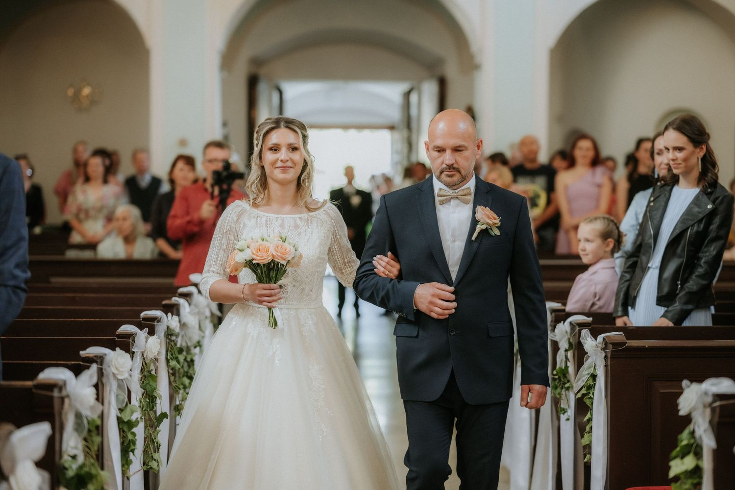Bride in white gown walks down church aisle with escort holding bouquet during wedding ceremony.