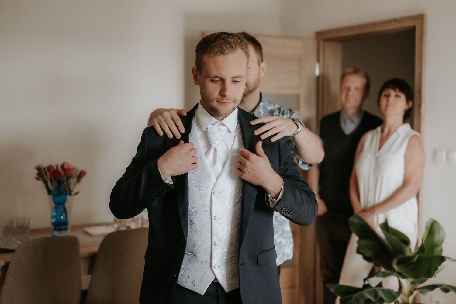 Groom preparing for wedding, adjusting suit jacket with help, family watching in background.