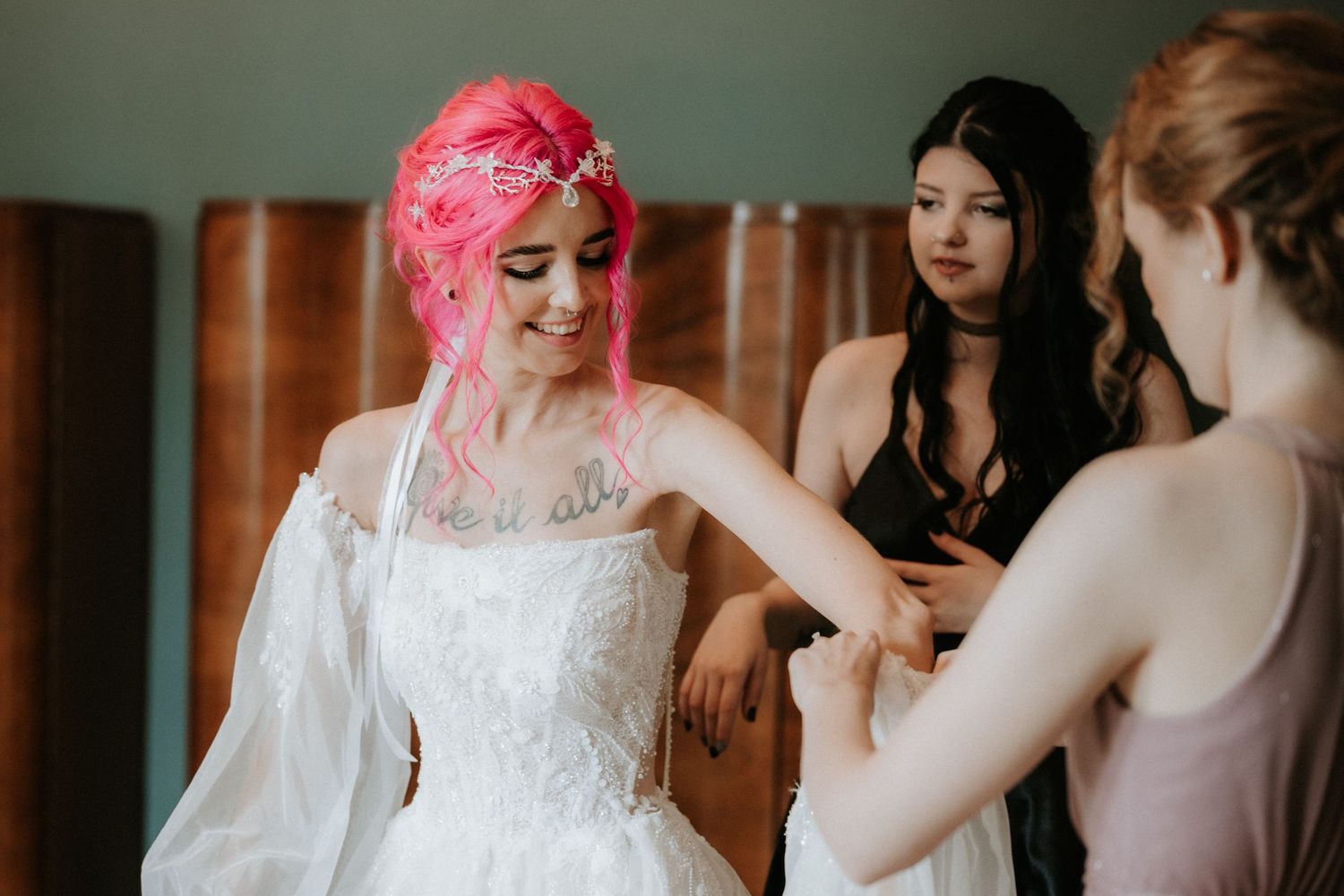 Bride with pink hair in white lace wedding gown being helped by bridesmaids during bridal preparation.