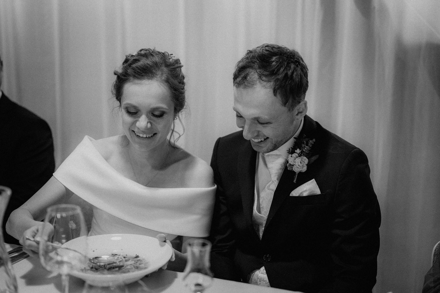 Black and white photo of a smiling bride and groom sitting together at their wedding reception dinner table.