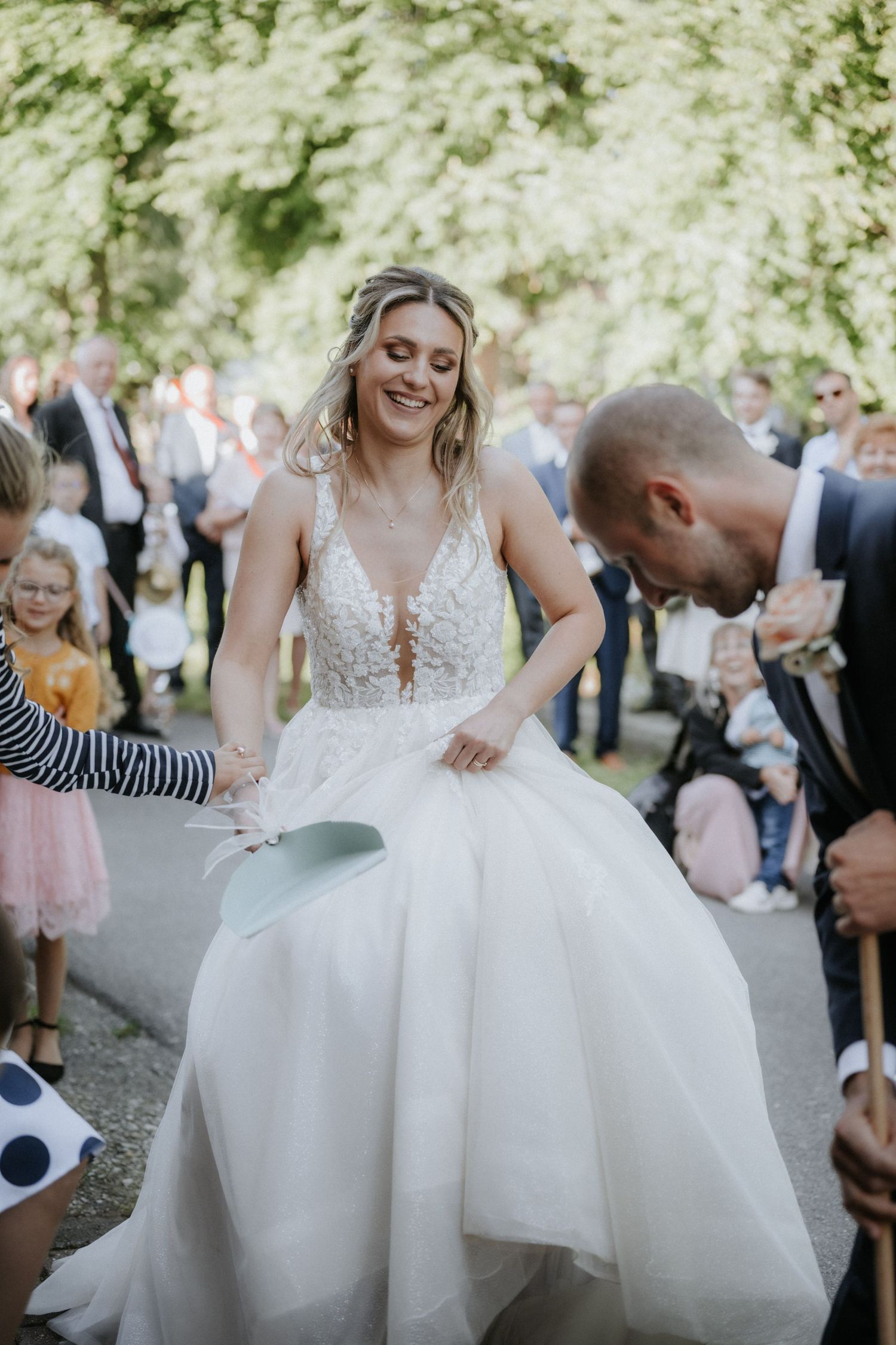 Bride in floral wedding gown laughing during outdoor ceremony while groom and guests celebrate around her.