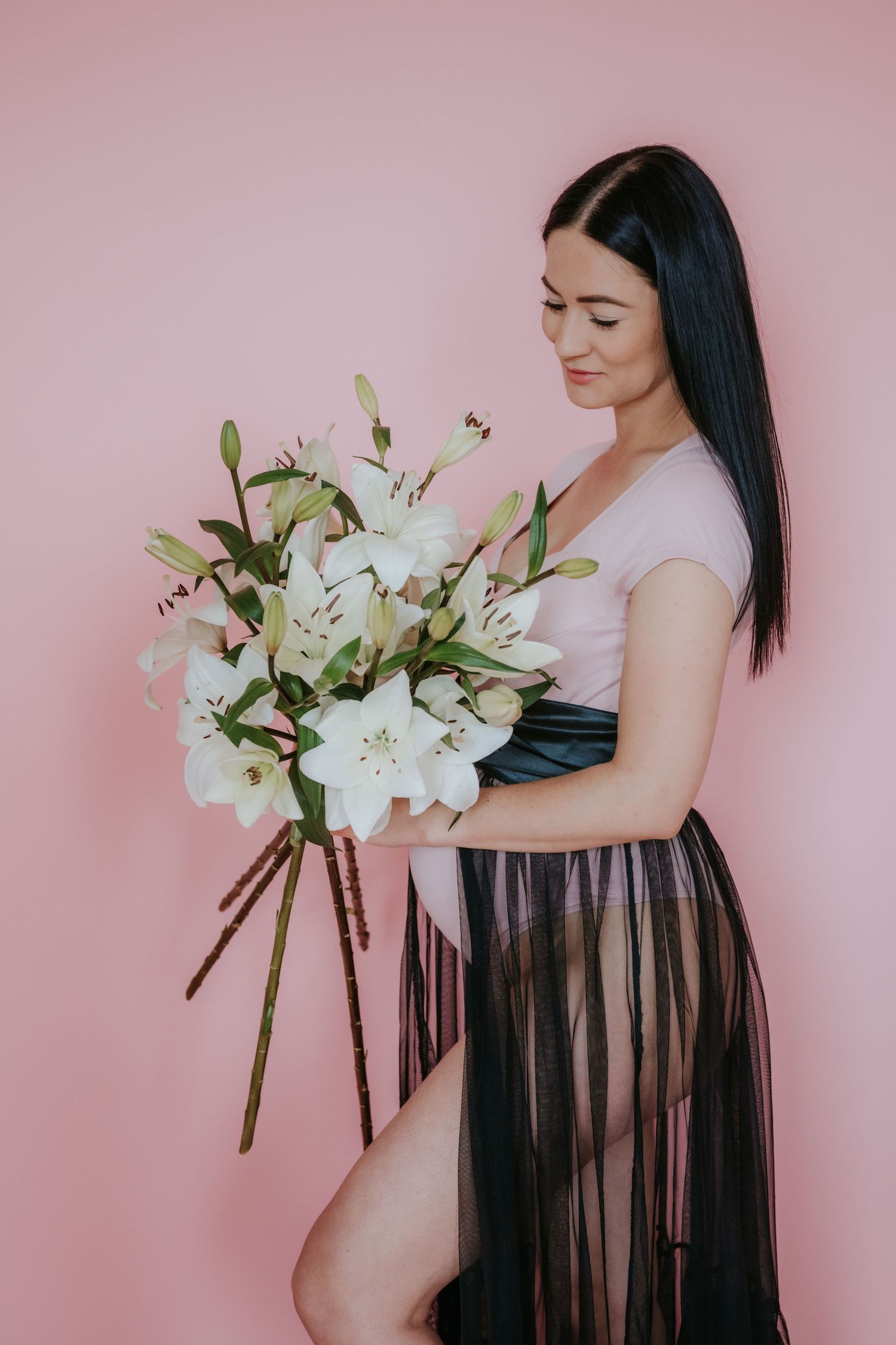 Brunette woman in pink bodysuit and black sheer skirt holding large white lily bouquet on pink background.