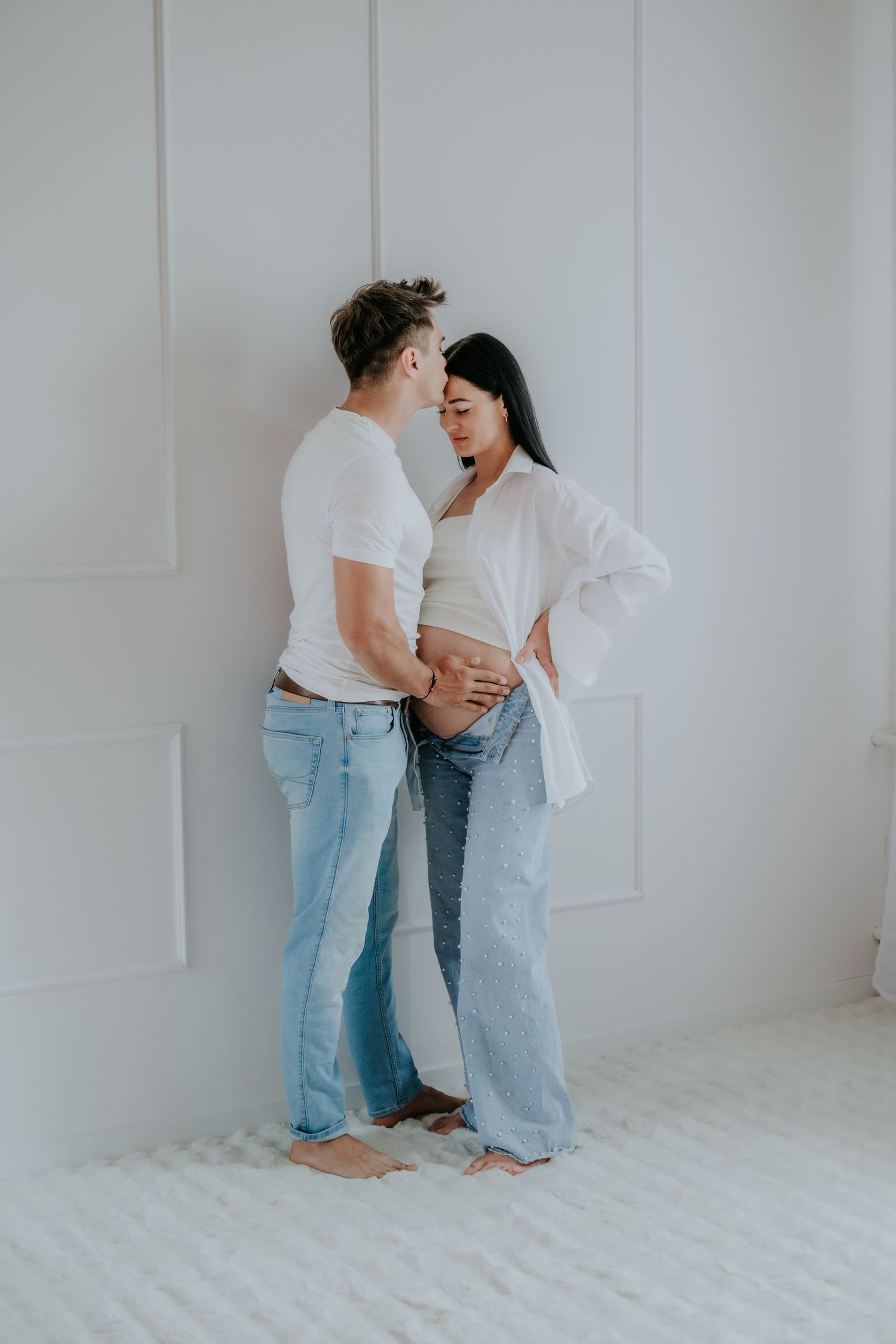 Couple in jeans embracing tenderly, mans hands on pregnant womans belly against white wall.