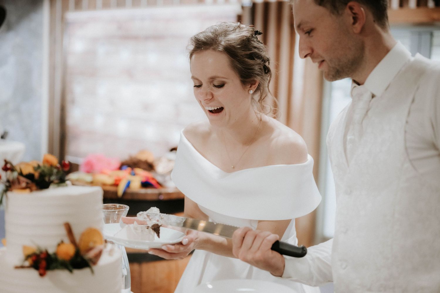 Bride and groom cutting their wedding cake together, bride in off-shoulder white gown, groom in white suit.