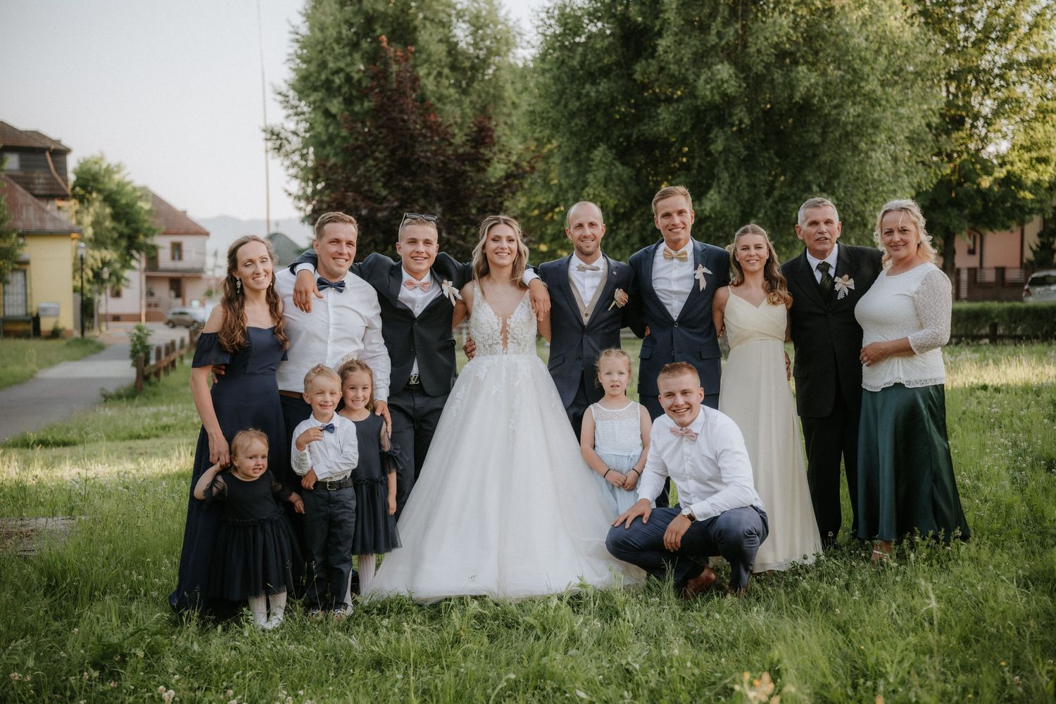 Wedding party group photo outdoors on green grass, bride in white gown surrounded by family in formal attire.