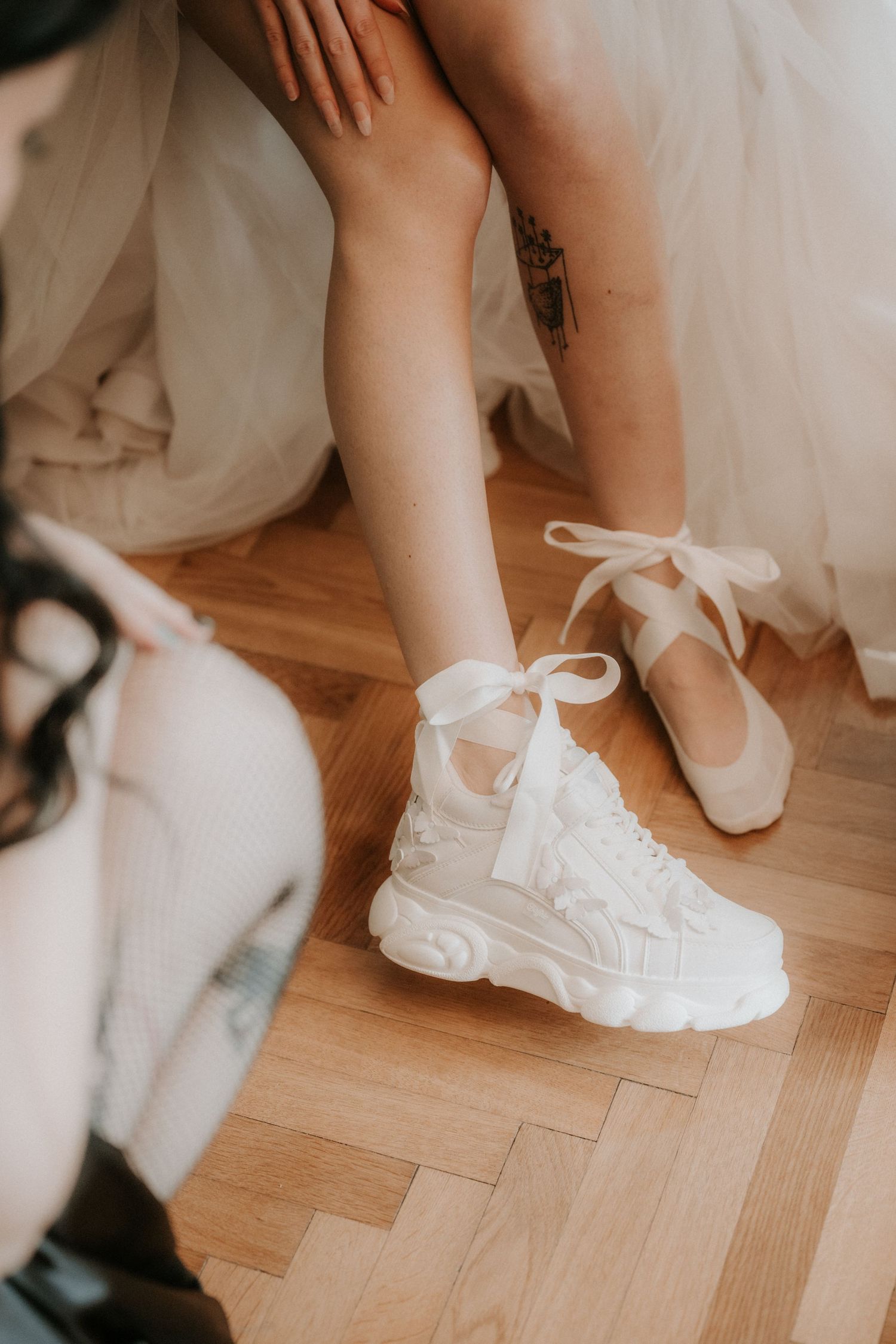 Bride wearing white chunky sneakers with ribbon laces beside ballet flats on wooden floor.