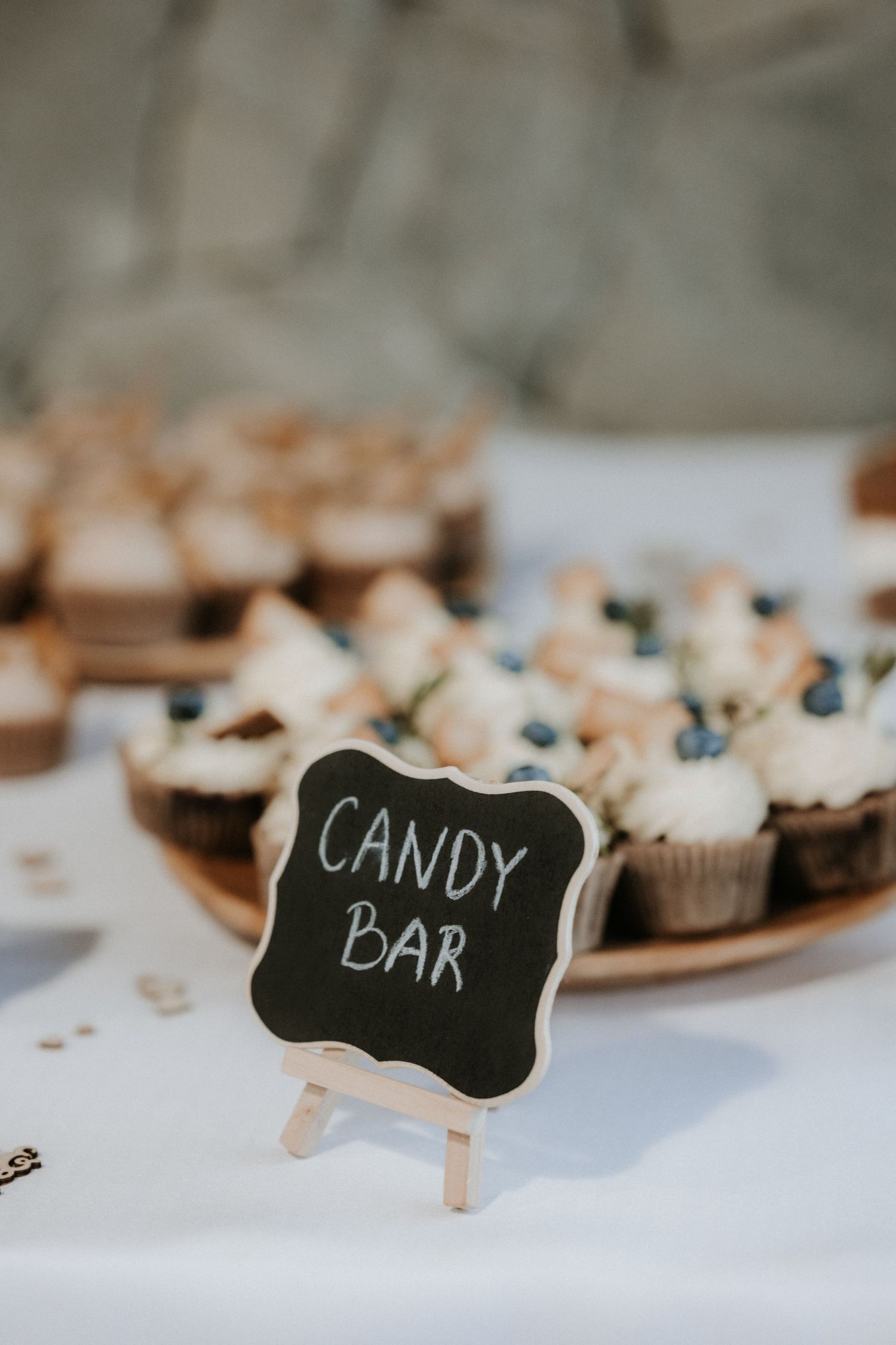 A chalkboard candy bar sign on a easel in front of cupcakes with white frosting and blueberries at a wedding.